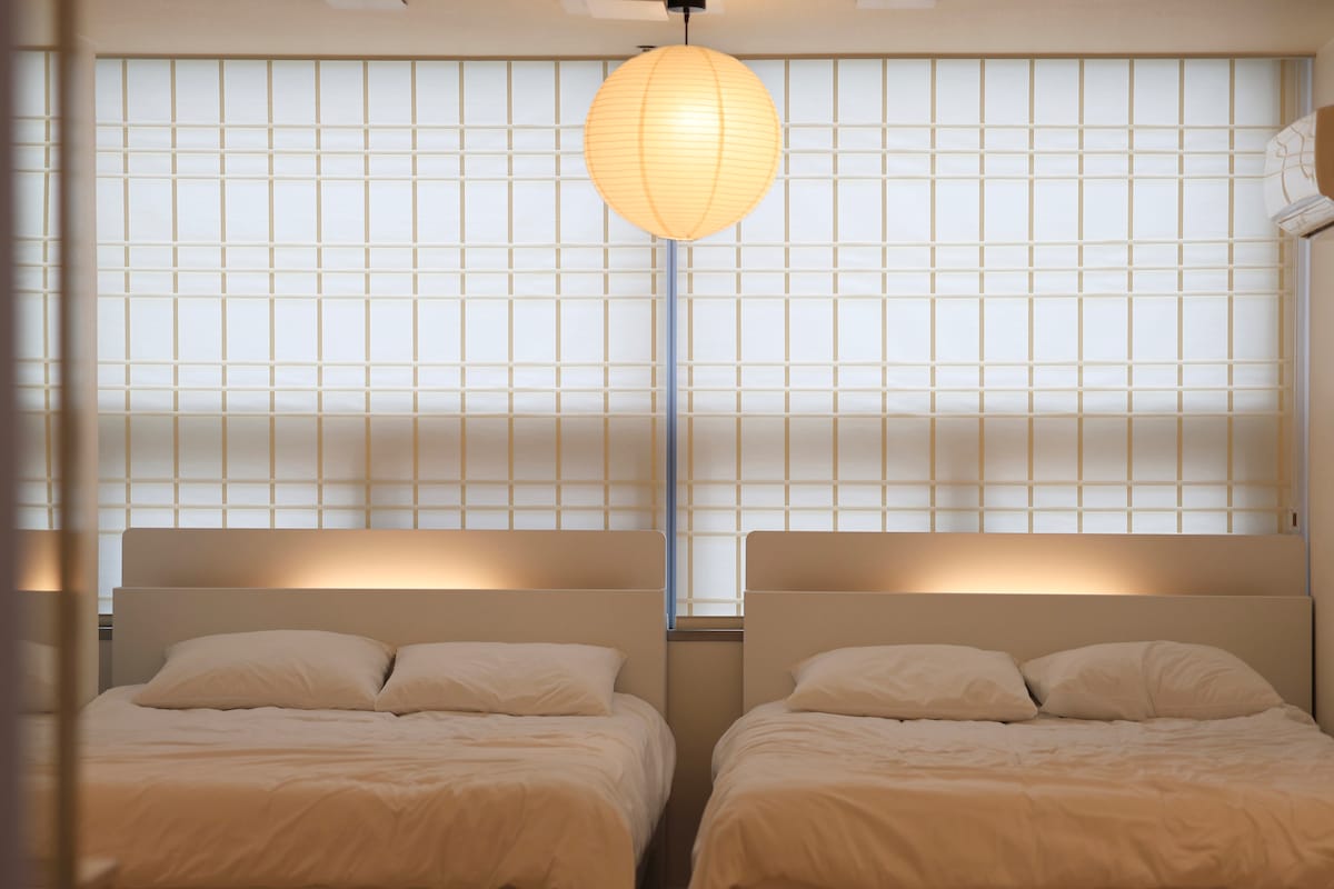 A well-lit bedroom features two spacious beds with comfortable pillows positioned beneath soft window blinds that filter natural light. A round paper lantern hangs from the ceiling, providing an ambient glow that enhances the serene atmosphere.