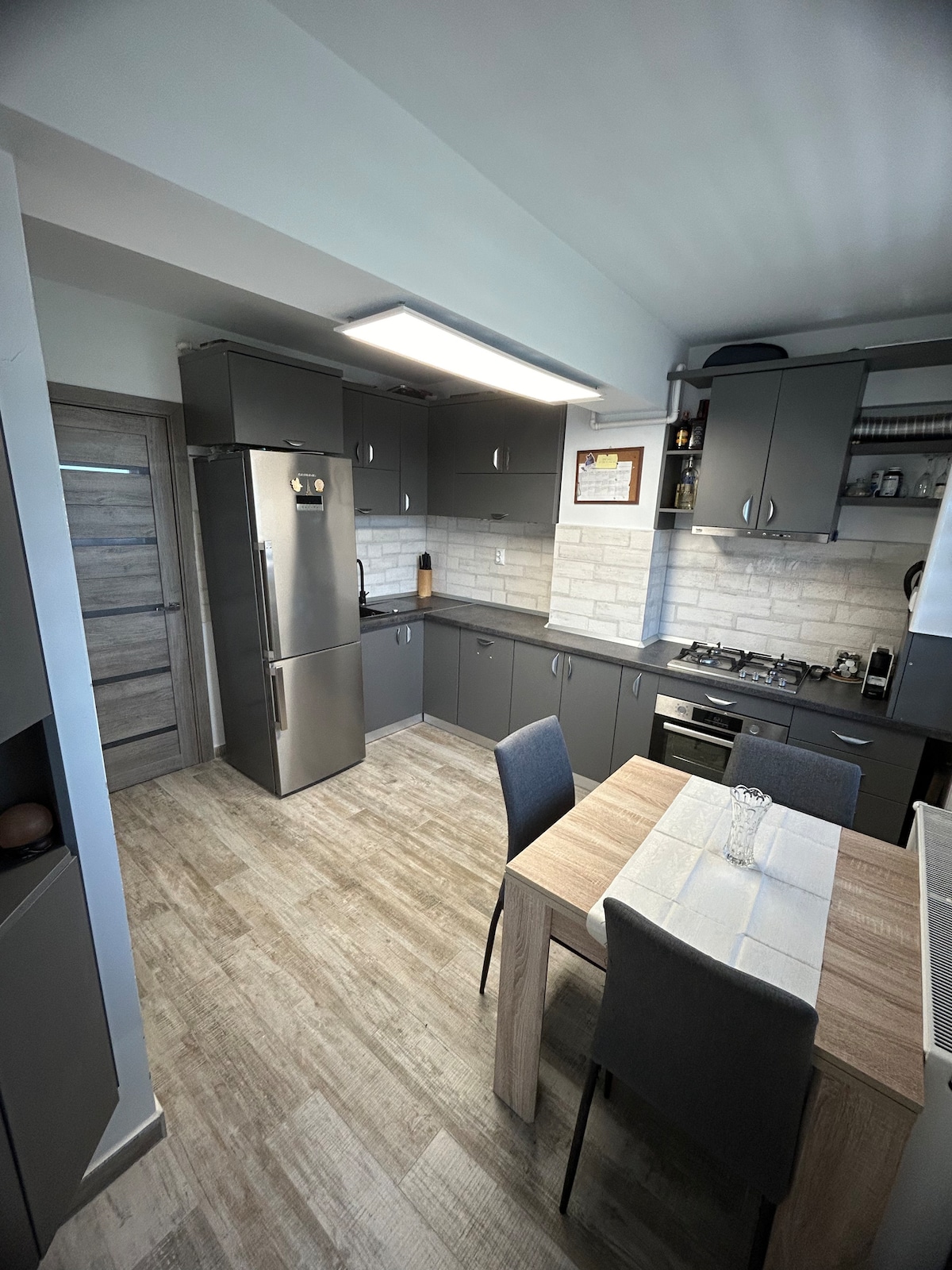 A modern kitchen featuring gray cabinetry and a stainless steel refrigerator. A wooden dining table is set with two gray chairs. Countertops are adorned with light-colored tiles, and appliances are arranged for convenience. Natural light illuminates the space from a nearby window.