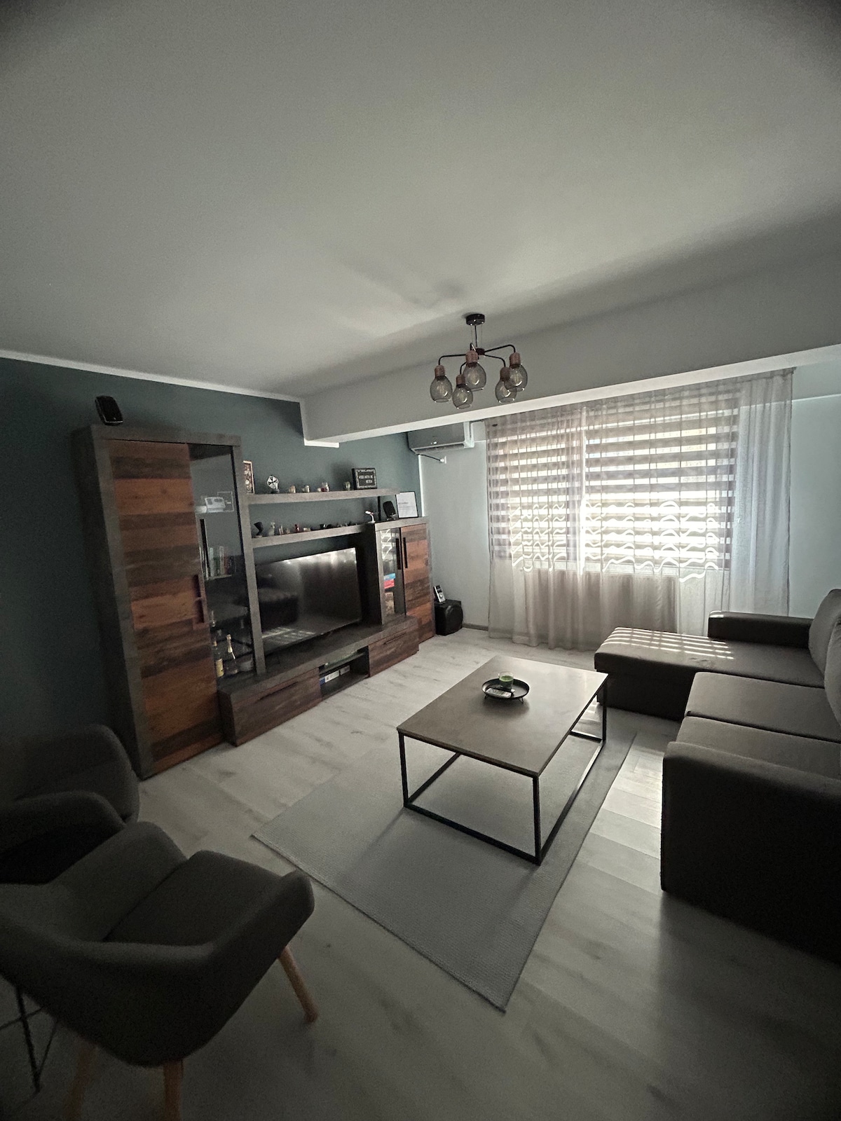 A modern living area features a comfortable sectional sofa and a wooden coffee table. A sleek TV unit is positioned against the wall, complemented by built-in shelving. Natural light filters through the sheer curtains, creating a calm, inviting space.