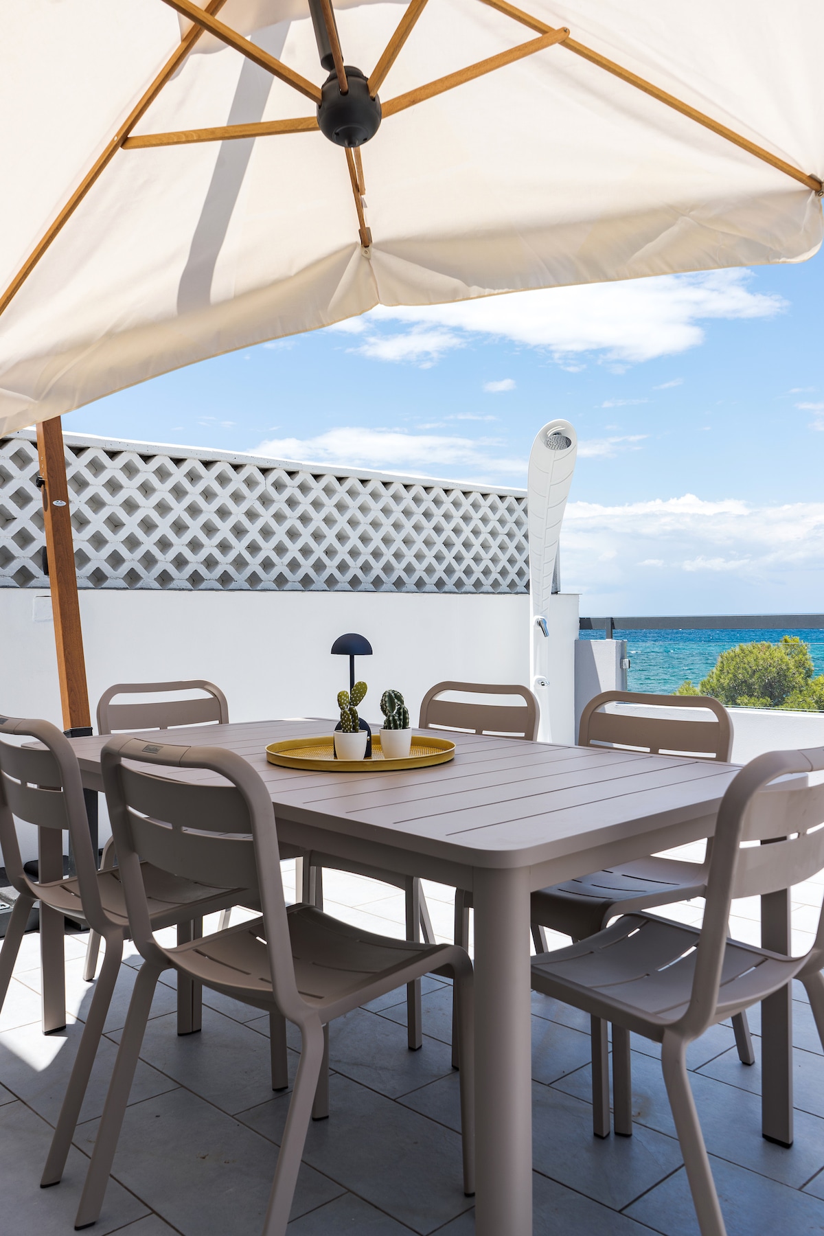 A dining area is set under a large umbrella, featuring a rectangular table surrounded by six chairs. A decorative tray with small plants is placed in the center. The view of the sea is visible in the background against a clear blue sky.