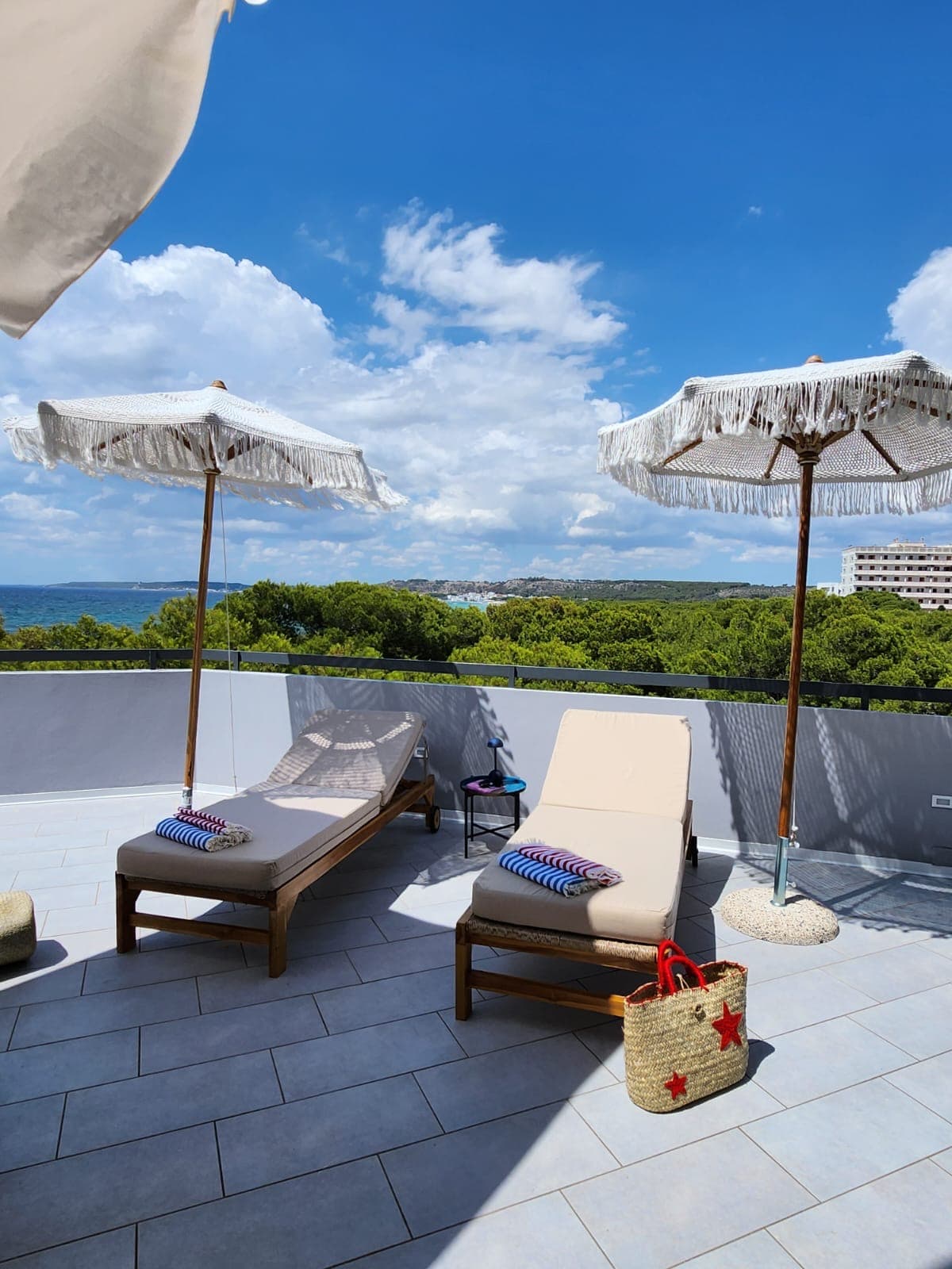 Two sun loungers are positioned on a tiled terrace, accompanied by large umbrellas providing shade. A view of the ocean and greenery is visible in the background, enhancing the outdoor space's inviting nature.