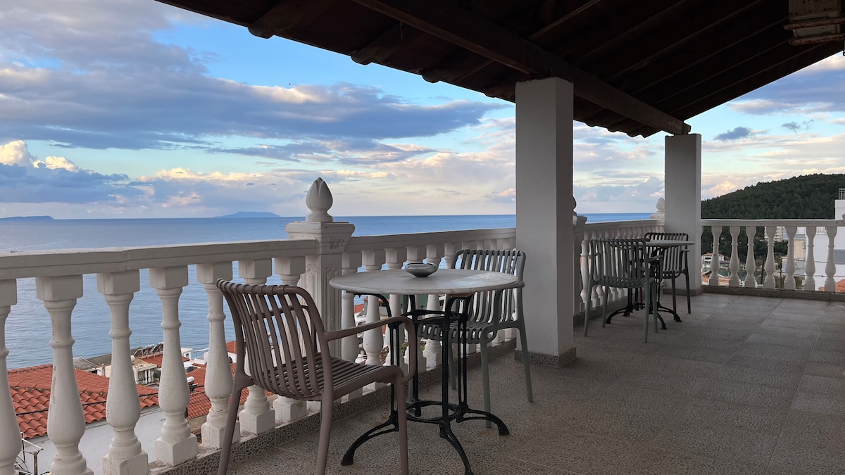 A spacious terrace offers a stunning view of the sea, with a mix of tables and chairs arranged for relaxation. The railing features ornate white balusters, enhancing the open feel. The sky is filled with clouds, creating a peaceful atmosphere for guests.