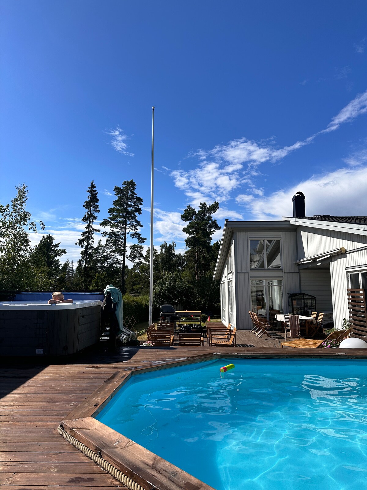 A wooden deck surrounds a sparkling blue swimming pool, with a spacious hot tub nearby. Sun loungers are positioned on the deck, offering a relaxing space. A tall flagpole stands against a backdrop of green trees and a clear blue sky, with clouds scattered above.