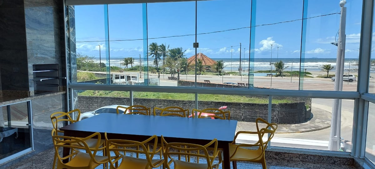 A dining area features a large table with bright yellow chairs. Floor-to-ceiling windows provide an unobstructed view of the beach, palm trees, and ocean. Natural light fills the space, enhancing the connection between indoor and outdoor settings.