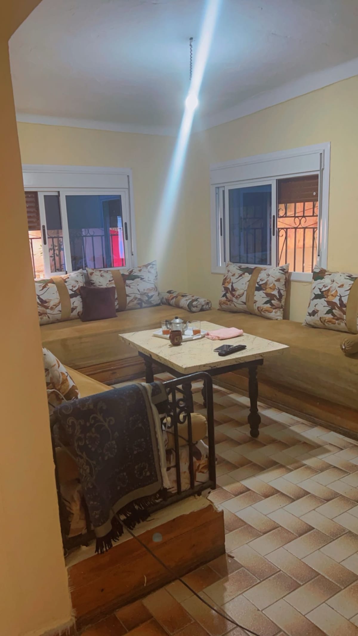 A welcoming seating area features a light-colored sofa adorned with floral cushions. A wooden coffee table occupies the center space, while the textured tile floor adds character. Large windows allow natural light to enter, enhancing the warm ambience of the room.