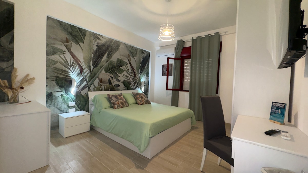 A spacious bedroom features a comfortable bed with a soft green coverlet, complemented by decorative pillows. Light wood flooring enhances the area. A flat-screen TV is mounted on the wall, while a white dresser and a dark chair are positioned nearby. Natural light is filtered through green curtains.