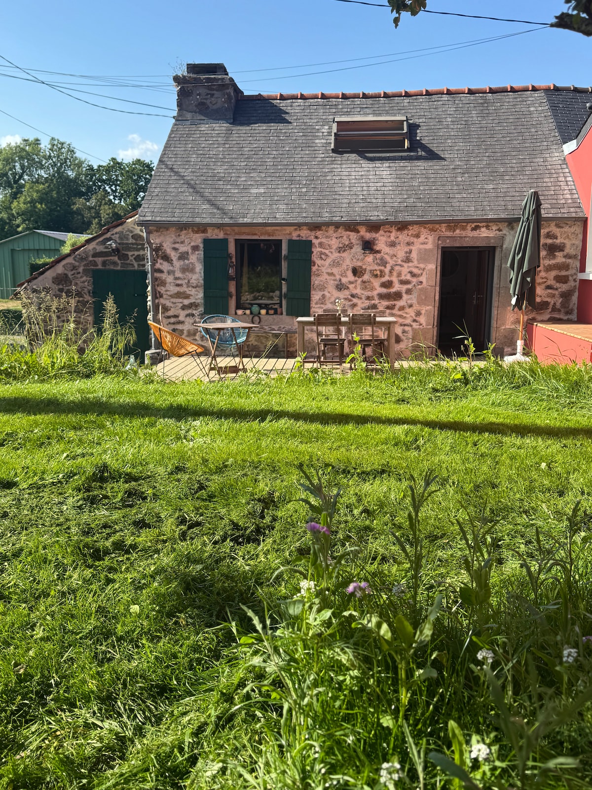 The exterior of the renovated cottage features a wooden terrace with a small dining table and chairs. Green shutters complement the stone facade, and tall grass surrounds the property. A parasol provides shade, enhancing the outdoor seating area.