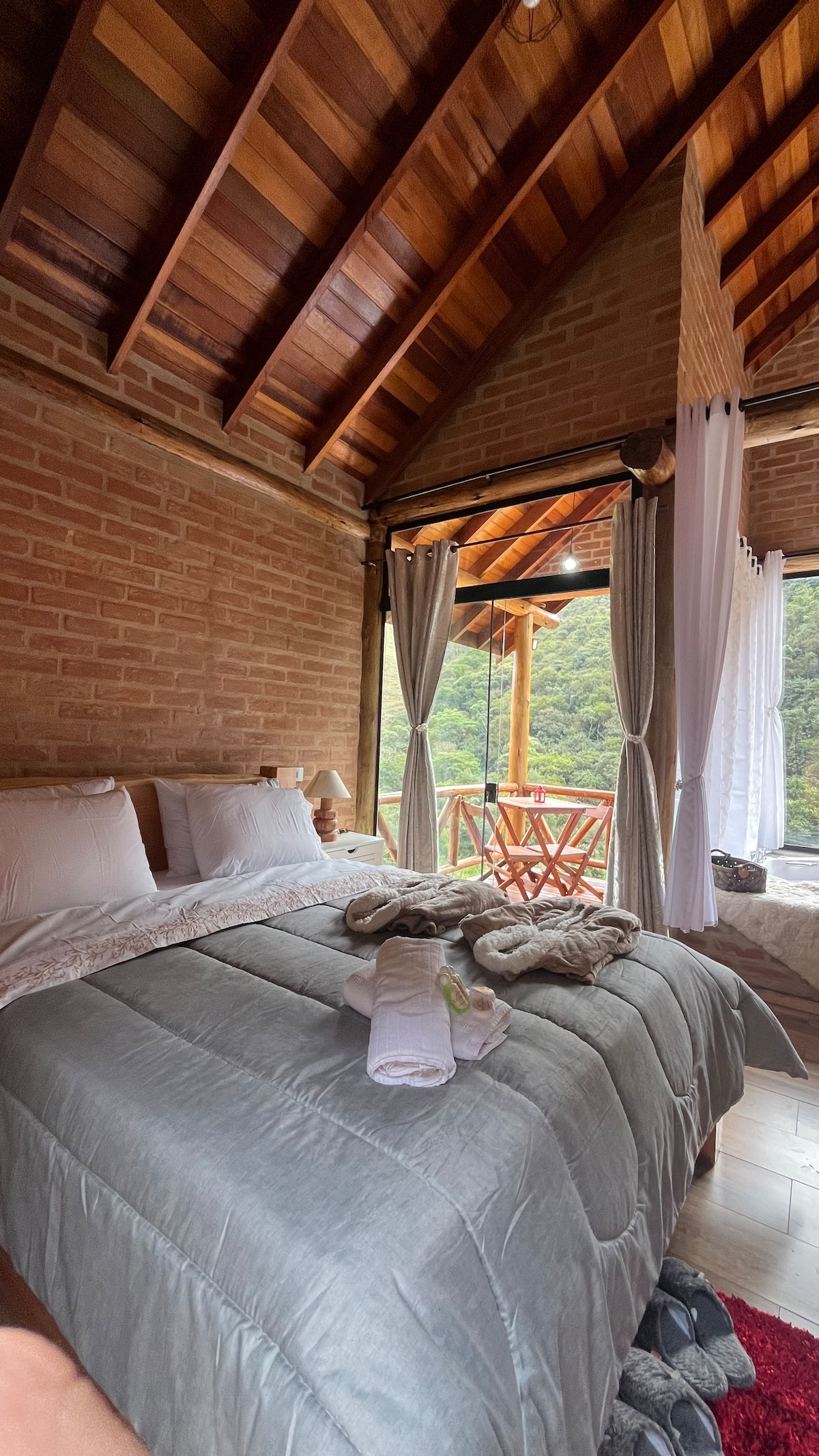 A cozy sleeping area features a spacious bed with light bedding, complemented by natural wood details and exposed brick walls. Large glass doors open to a private balcony with a small table and chairs, offering views of the surrounding nature.