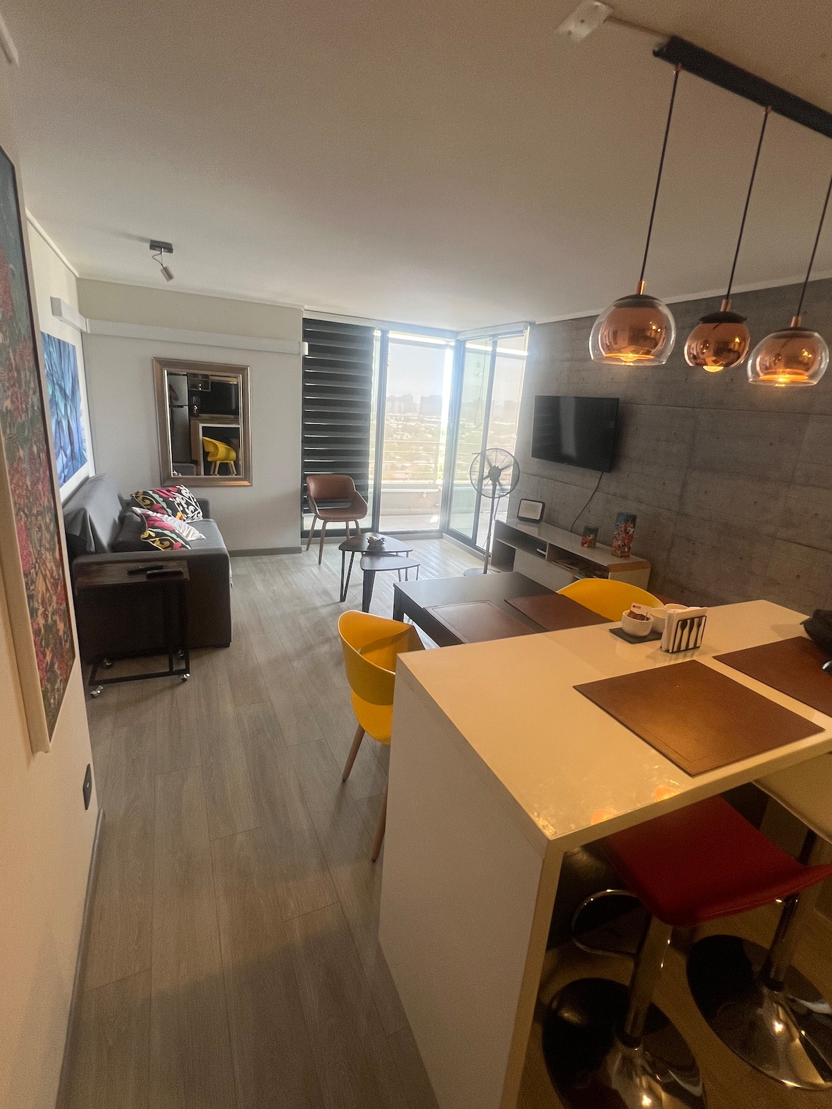 A modern living space features a sleek design with a concrete wall and ample natural light from the large windows. A dining table is complemented by colorful chairs, while a cozy seating area includes a sofa and decor. An entertainment unit and fan are also visible.