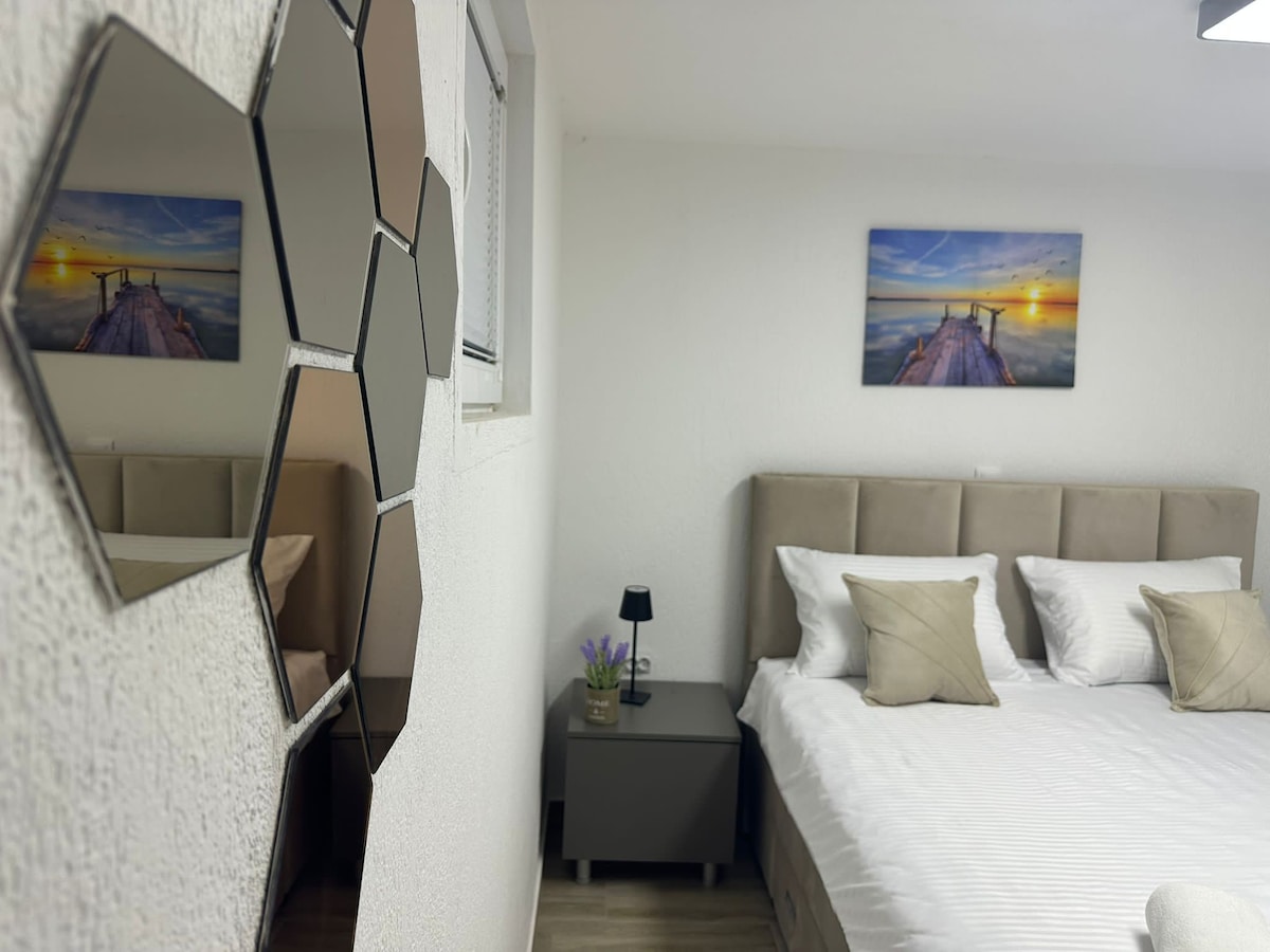 A bright sleeping area is featured, showcasing a spacious bed with light-colored linens and accent pillows. A bedside table holds a small lamp and a floral arrangement. Mirrors in a geometric shape decorate the wall, reflecting the serene artwork above the bed.