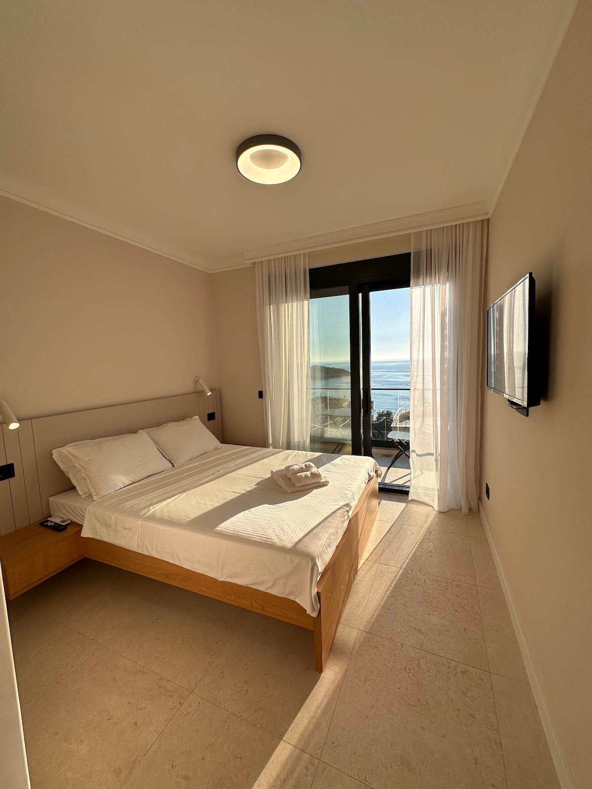 A light-filled bedroom features a spacious bed with fresh linens, framed by large glass doors that lead to a private balcony with a sea view. Soft curtains allow natural light to brighten the room, creating a calm and inviting atmosphere.