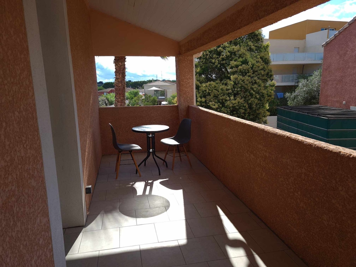 A private terrace is showcased, featuring a round table and two chairs. Sunlight creates a pattern on the tiled flooring, while the surrounding walls provide a sense of enclosure. Views of greenery and nearby structures are visible beyond the railing.
