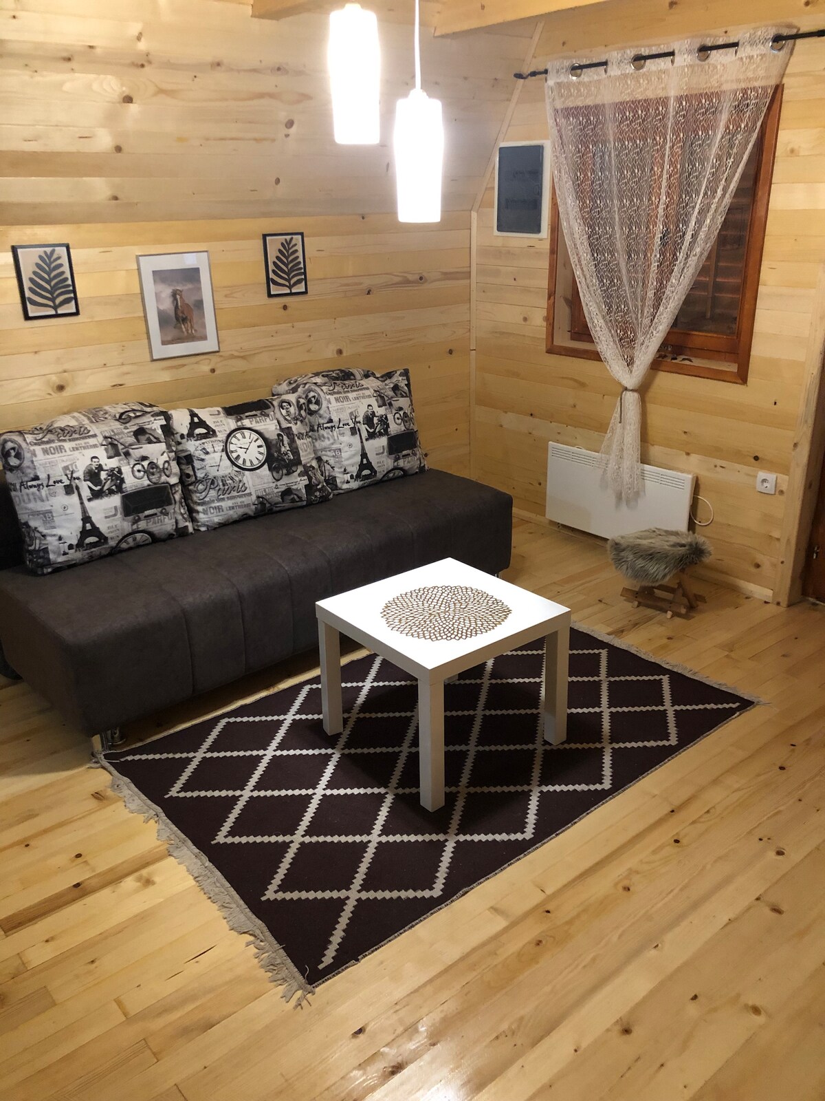 A cozy living area is highlighted by wooden walls and warm lighting. A dark sofa with decorative cushions faces a small white coffee table, while a patterned rug lies beneath. A sheer curtain frames a window, enhancing the inviting ambiance.
