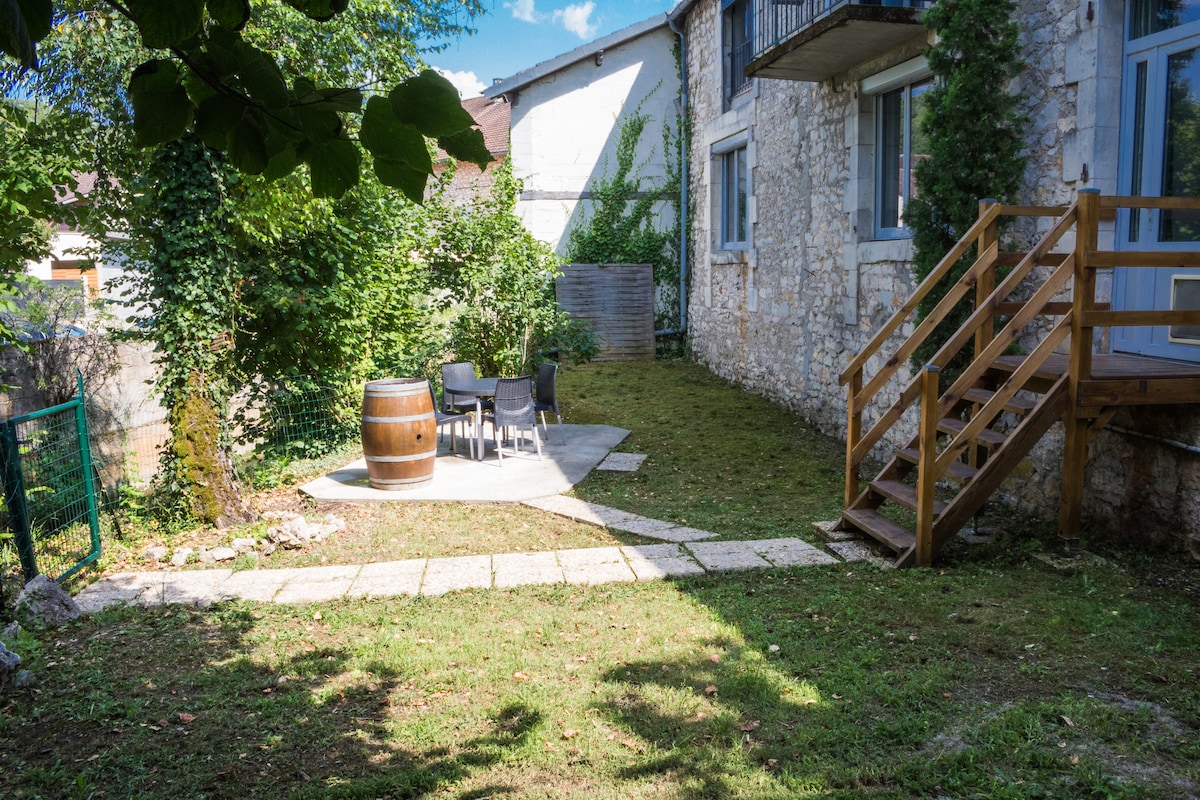 The outdoor space features a private garden with a neatly manicured lawn, a stone patio, and a wooden barrel surrounded by four chairs. Direct access is provided through wooden steps from the apartment. Lush greenery adds a natural element to the serene setting.