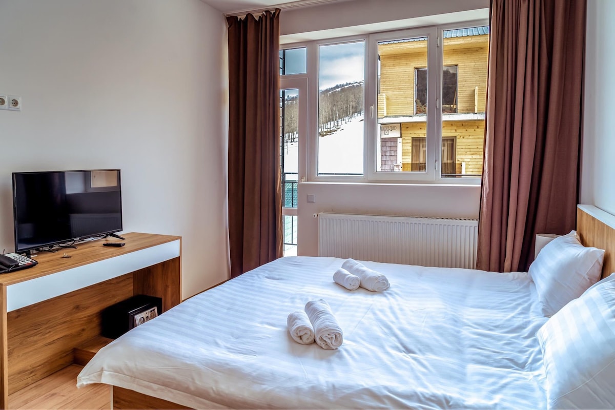 A well-lit bedroom features a spacious bed with crisp white linens. Two neatly rolled towels are placed on the bed. A small television is positioned on a wooden desk, and large windows showcase views of the surrounding landscape.