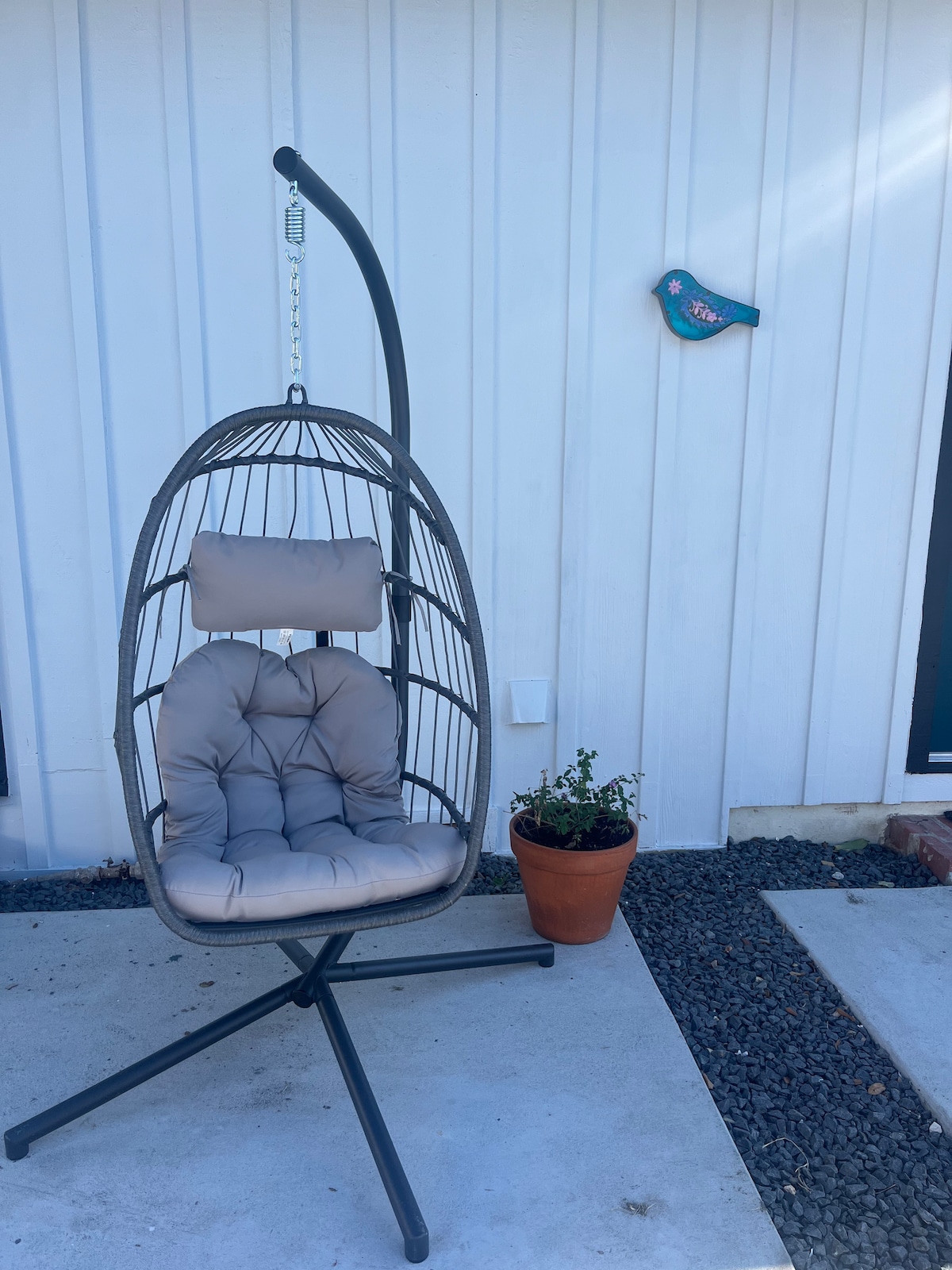 A unique hanging chair is situated on a concrete patio. It features a cushioned seat and backrest within a woven frame. A small potted plant is positioned nearby, adding a touch of greenery, while a decorative bird is mounted on the white wall.