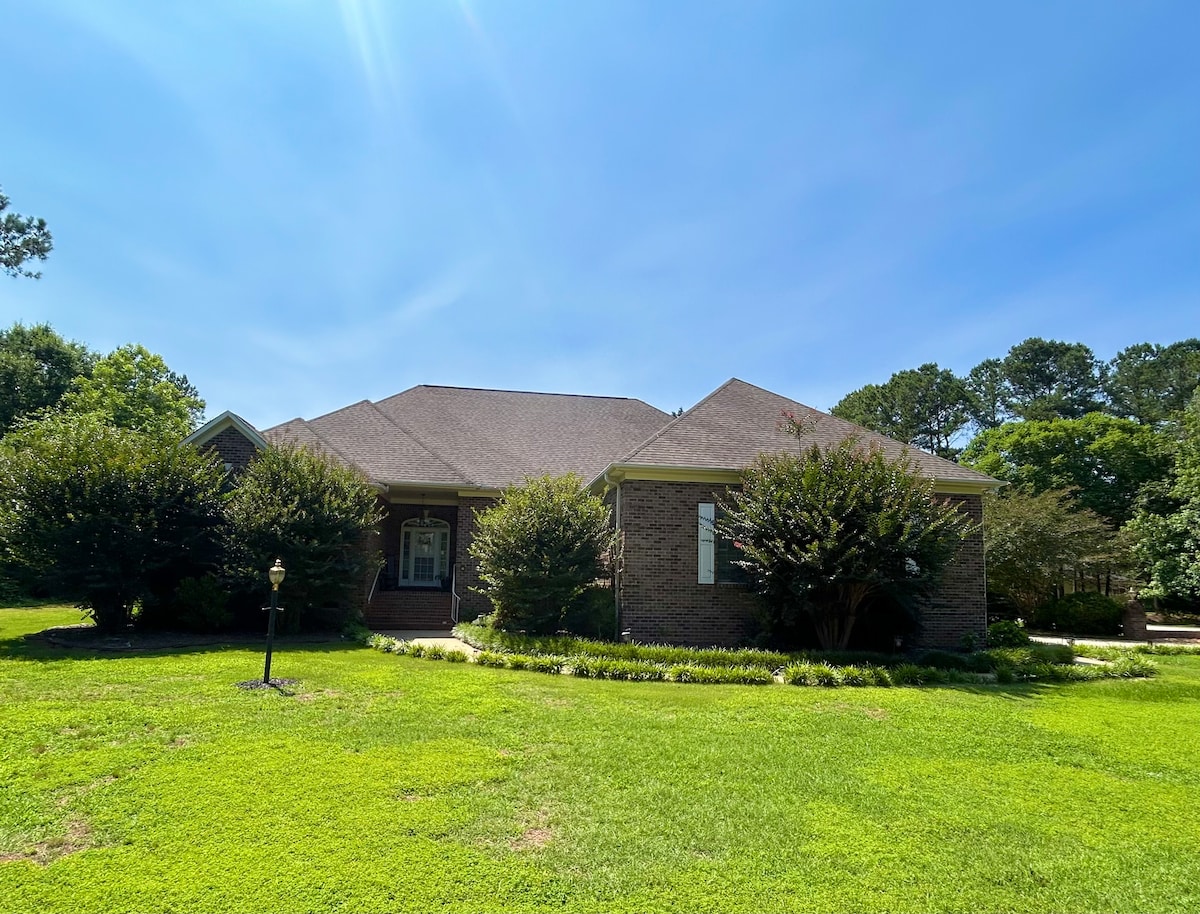 A single-story brick home is surrounded by lush green grass and well-maintained shrubbery. The structure features a broad roofline and multiple windows, providing a welcoming appearance. A lamppost stands in the front yard, contributing to the property's charm.