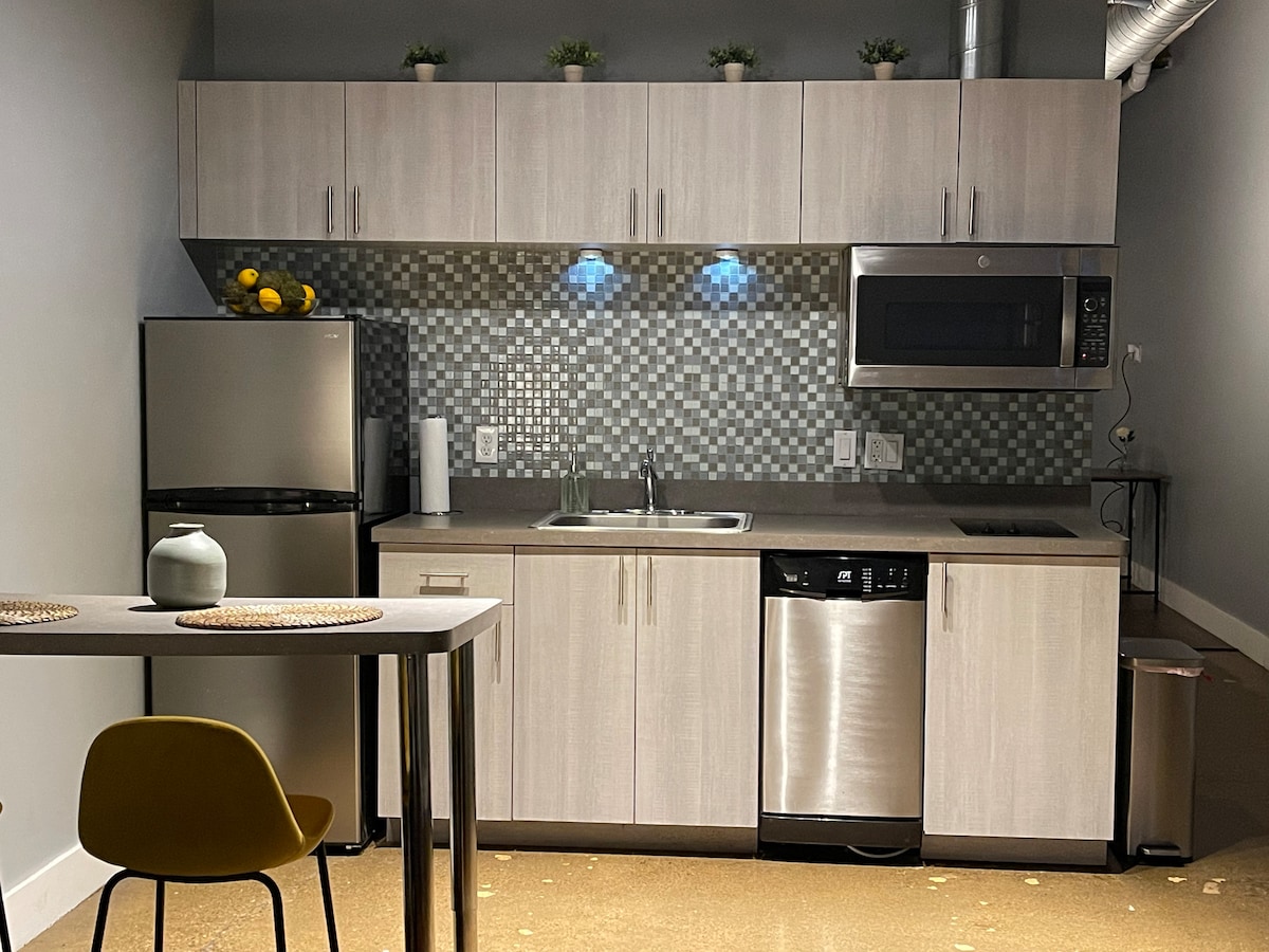 A modern kitchen is equipped with stainless steel appliances, including a microwave and refrigerator. Luminous blue accent lighting highlights the mosaic tile backsplash. A small dining table with a yellow chair is positioned nearby, creating a functional and inviting cooking area.