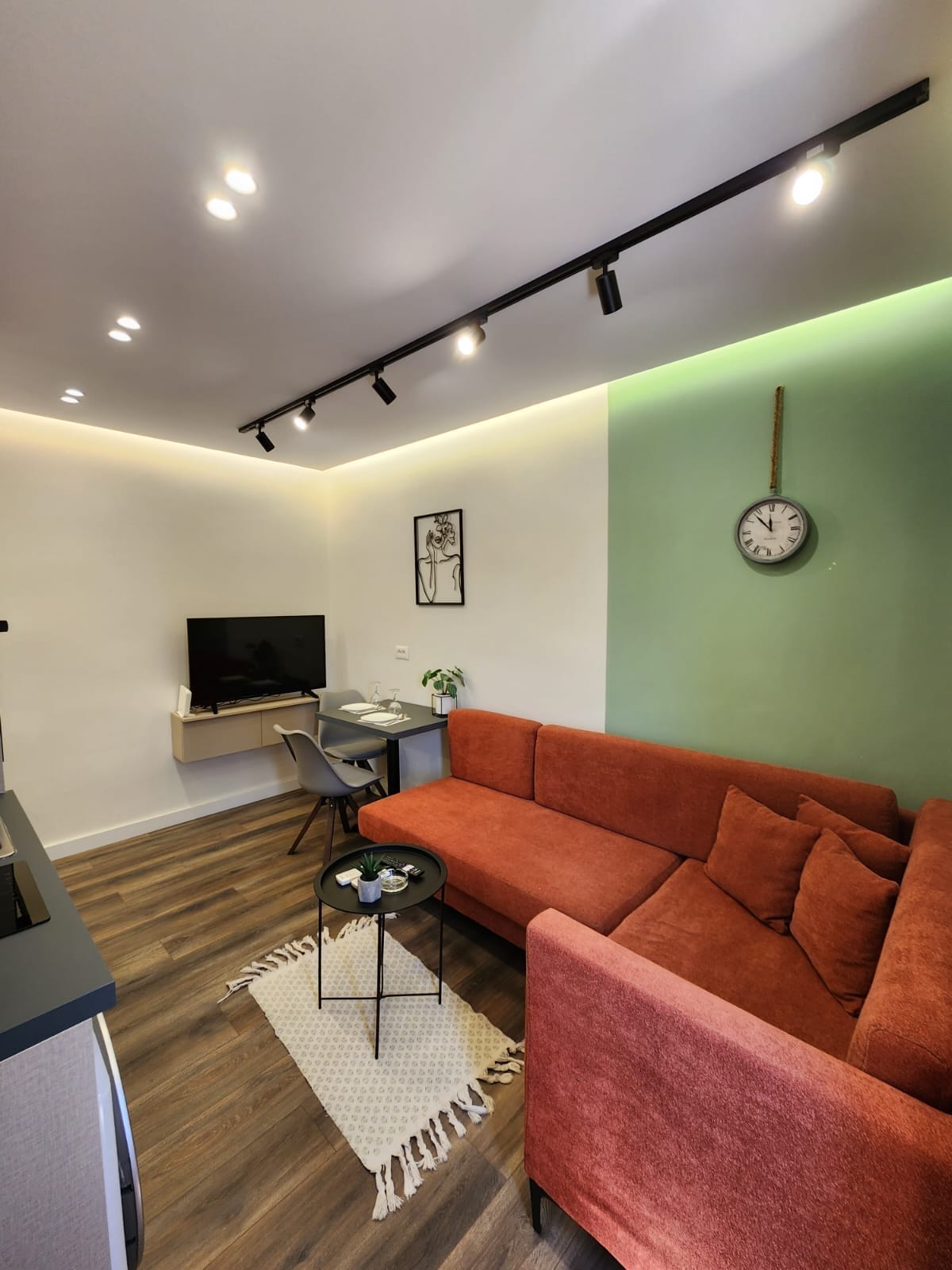 A cozy living area features a vibrant orange sectional sofa, complemented by a small black table and modern desk. A wall clock adds a stylish touch above the sofa, while soft lighting enhances the space. A TV is mounted on the wall, providing entertainment options.
