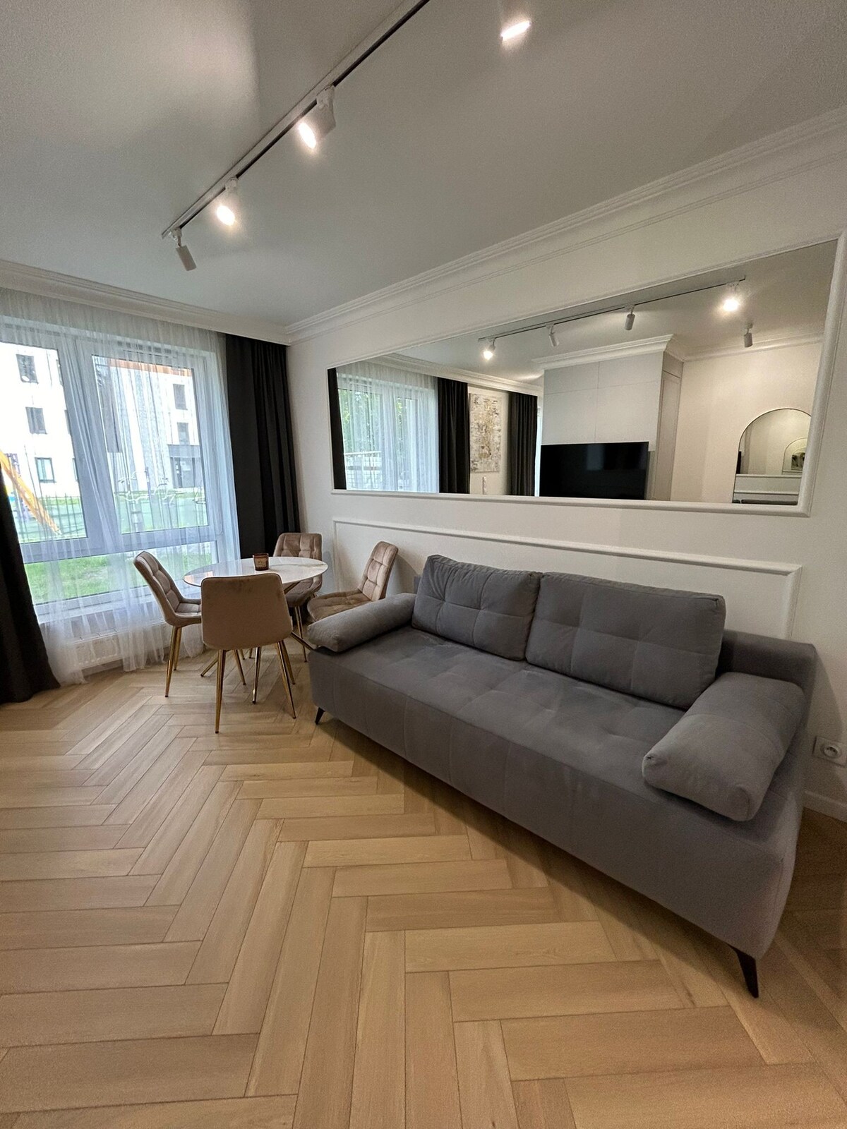 A modern living area features a gray sofa with soft cushions and a stylish dining table set for four. Large windows allow natural light to fill the room, while light wood flooring adds a warm touch to the space.
