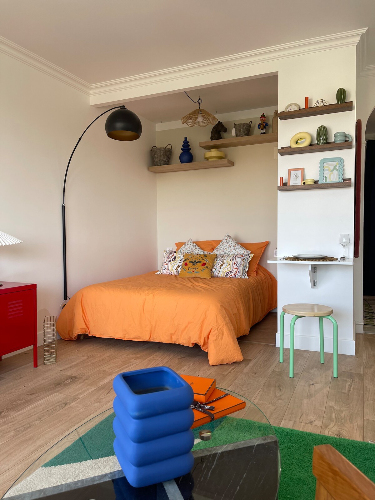 A vibrant sleeping area features an orange bedspread complemented by decorative pillows. A stylish arc lamp stands nearby. Additional shelves display various artistic objects, while a small table and stool create a cozy seating space, highlighting the studio's modern and colorful design.