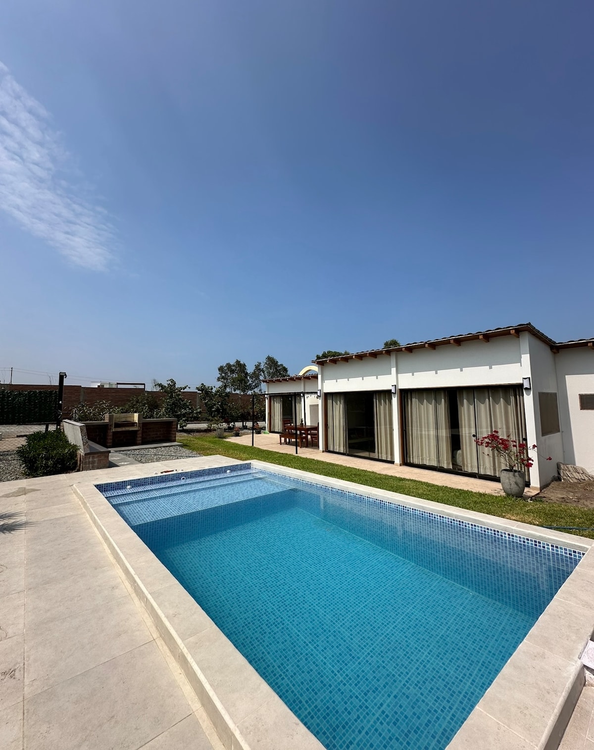 A private pool glistens under a clear blue sky, surrounded by a patio area. A ranch-style house with large windows and sliding doors is visible, providing a view of the lush, green garden. The space offers relaxation and opportunities for outdoor enjoyment.