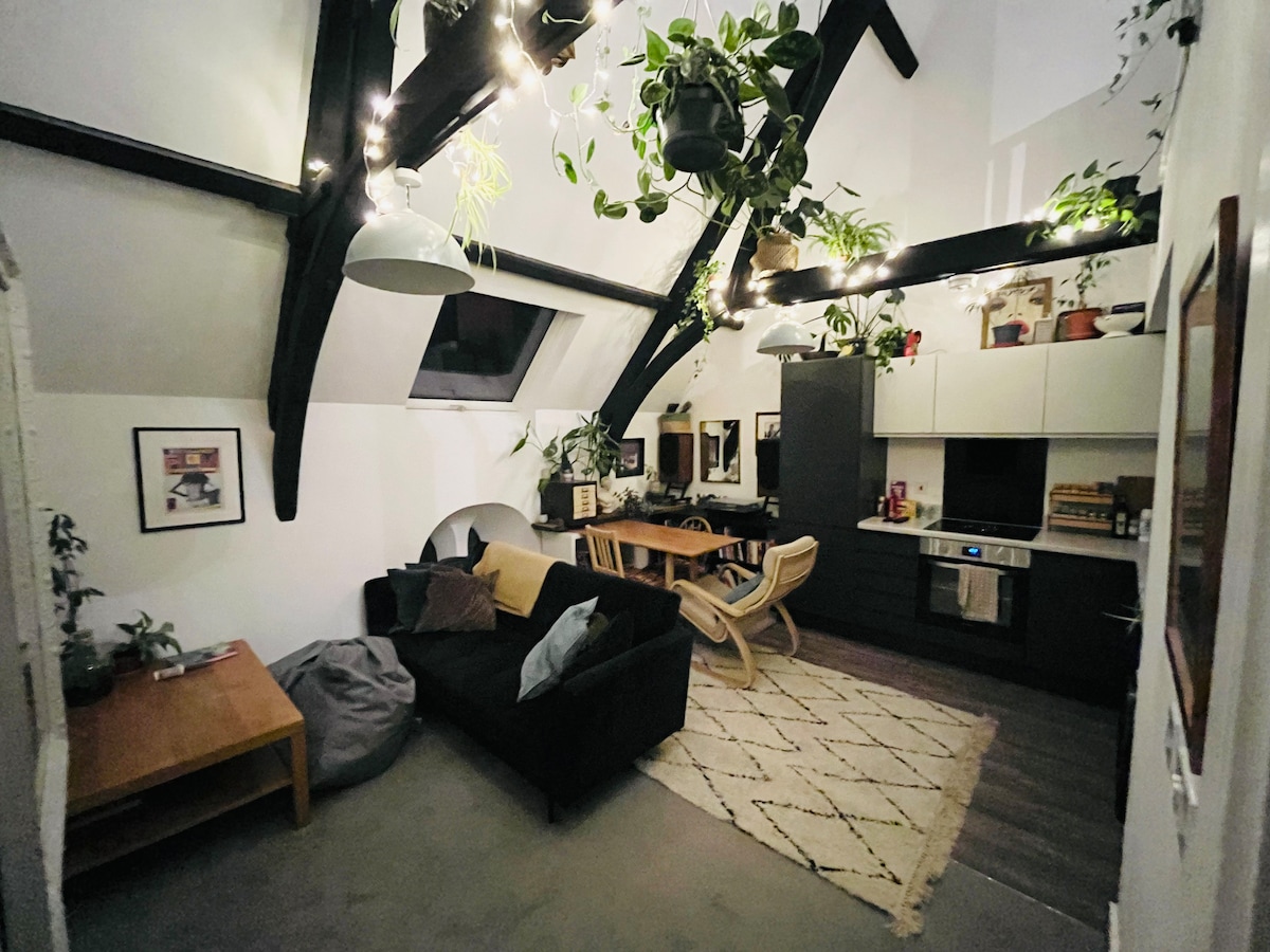An open living space features a dark sofa and a wooden dining table accompanied by stylish chairs. Ceiling beams and greenery are accentuated by string lights, creating a cozy ambiance. A well-equipped kitchen area is visible in the background, enhancing the overall functional design.