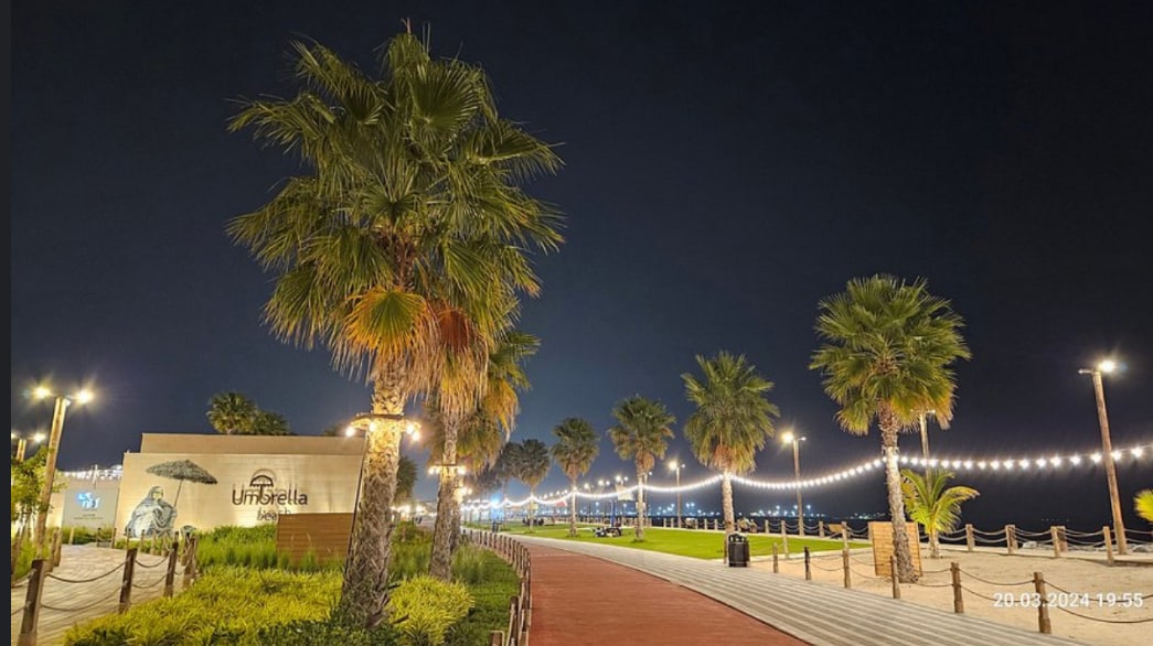 A walkway lined with palm trees is seen at night, illuminated by string lights that create a warm ambiance. The path is bordered by grassy areas and features a building with a sign, enhancing the inviting atmosphere of the beachfront promenade.
