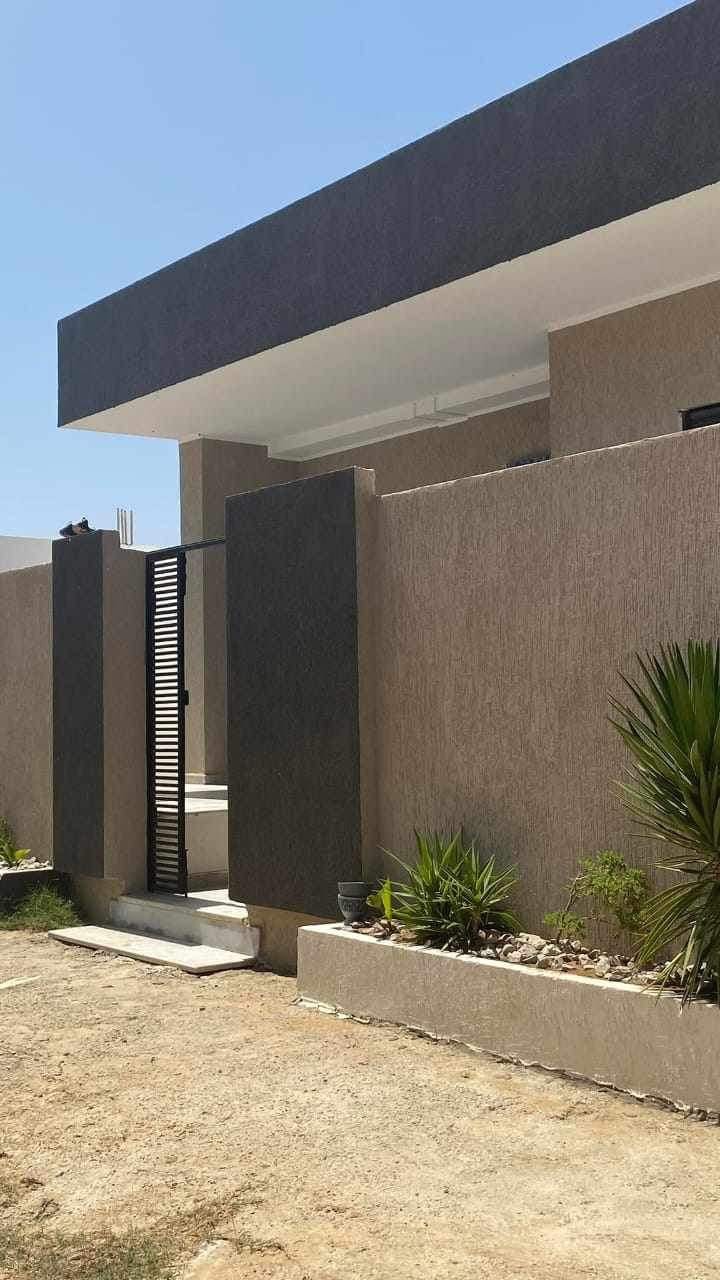 A modern exterior structure is presented, featuring neutral tones and sleek lines. Vertical slats are visible on the gate, flanked by planters with greenery. The entrance is enhanced by a small set of steps leading up to a doorway, surrounded by a well-maintained landscape.