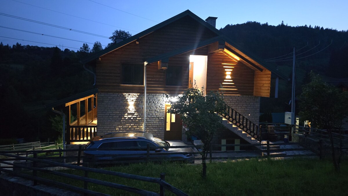 A charming house is depicted in the evening, featuring a mix of wooden and brick elements. Soft lighting highlights the facade, while surrounding trees and a grassy area create a serene setting. A car is parked in the driveway, adding to the accessible atmosphere.