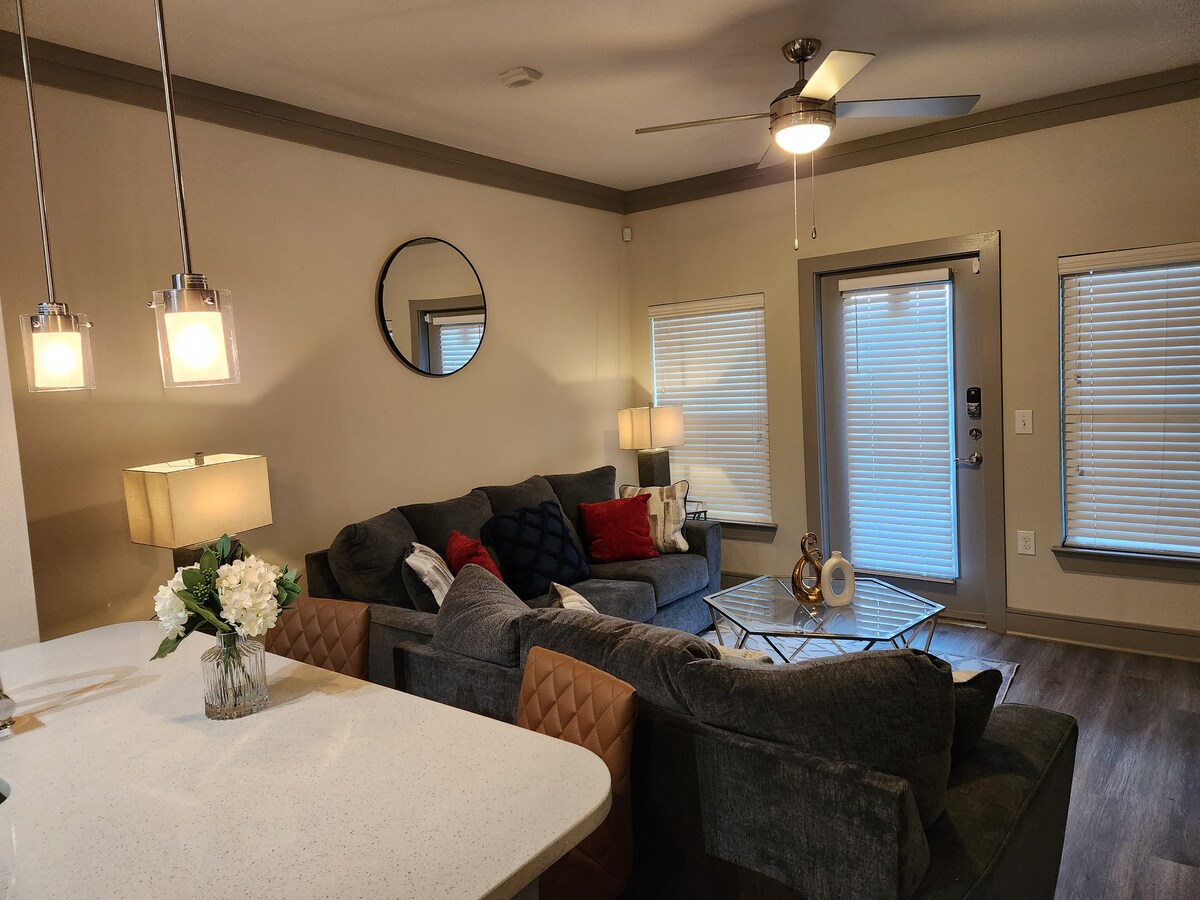 A modern living area features a large, plush sectional sofa adorned with decorative pillows. A glass coffee table sits at the center, surrounded by soft lighting from table lamps. Large windows with blinds allow for natural light, complementing the sleek design.