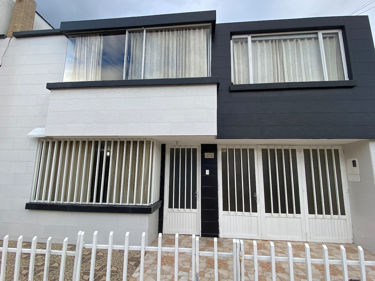 The exterior of the house presents a modern design with a two-tone color scheme of white and black. Large windows with sheer curtains allow natural light to enter. A gated entry leads to a tiled pathway, bordered by a white fence.