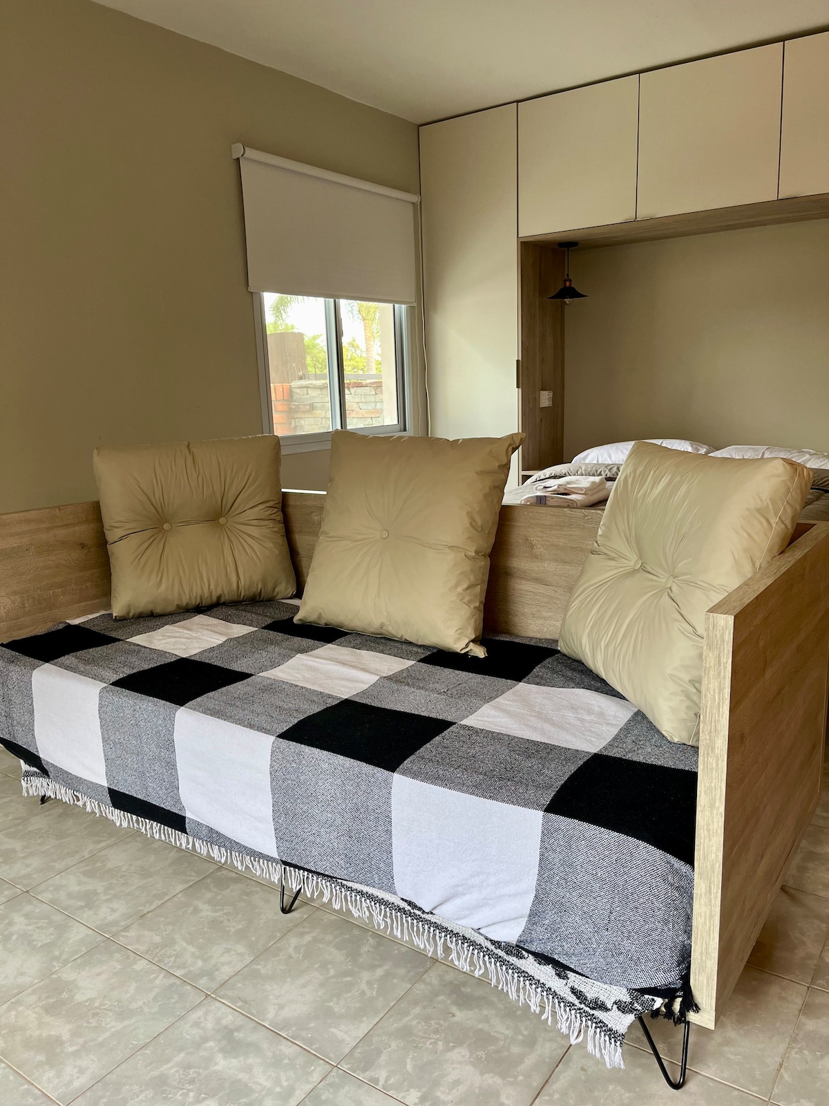 A cozy sofa bed is displayed in a neutral-toned room. The sofa features two plush back cushions and is covered with a checkered blanket. Natural light enters through a window, providing a bright and inviting atmosphere. Light-colored cabinetry is visible in the background.