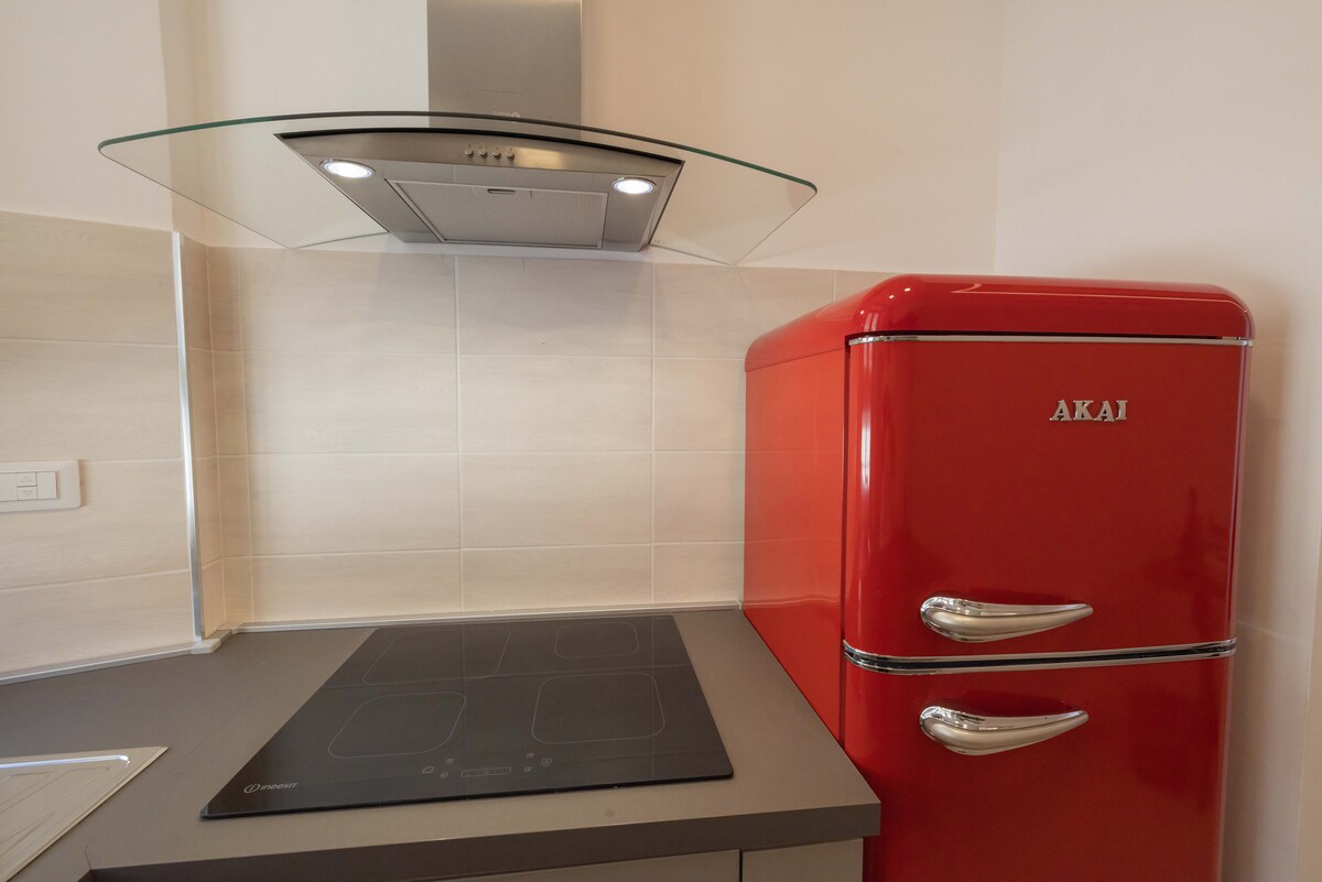 A modern kitchen space features a sleek, gray countertop with a cooktop and a striking red retro-style refrigerator. An extractor hood is positioned above, providing a functional addition to the area. Light neutral tiles line the walls, enhancing the contemporary design.
