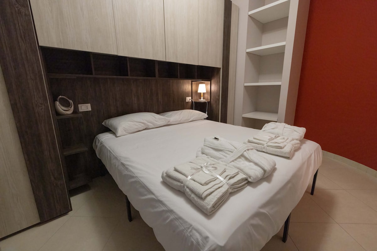 A neatly arranged sleeping area is highlighted by a spacious bed with fresh linens. Towels are folded at the foot of the bed, and a softly glowing lamp provides warm lighting. The walls feature a striking red accent, complemented by minimalist wooden storage.