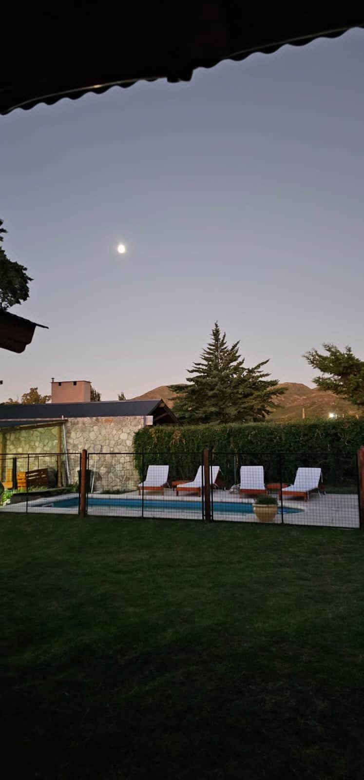 A serene outdoor space features a fenced pool area surrounded by well-maintained grass. Two lounge chairs are positioned beside the pool, with trees and distant mountains visible in the background under a clear evening sky.