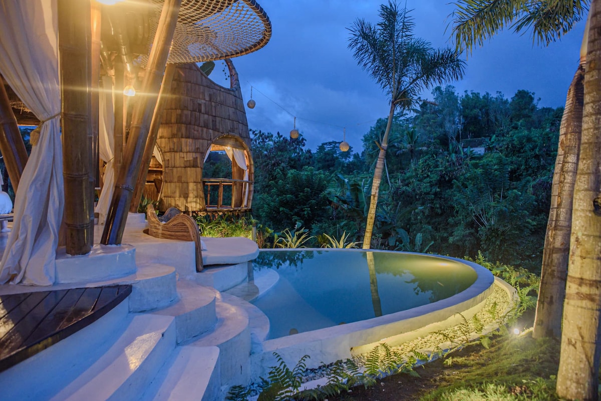 An outdoor area features a modern infinity-style pool surrounded by lush greenery. Steps lead from a bamboo structure to the pool, with gently curving edges enhancing the natural landscape. Soft lighting from nearby fixtures creates a serene ambiance in the evening setting.
