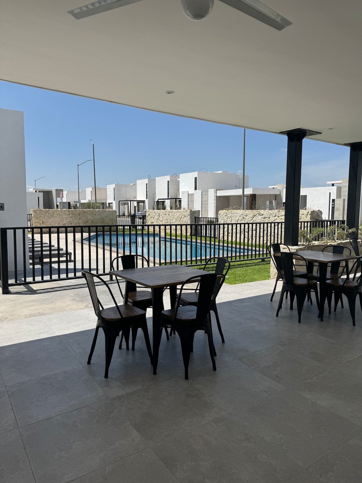 An outdoor dining area features several metal chairs and tables, positioned under a covered porch. A clear view of a swimming pool can be seen in the background, surrounded by landscaped grass and modern white buildings.