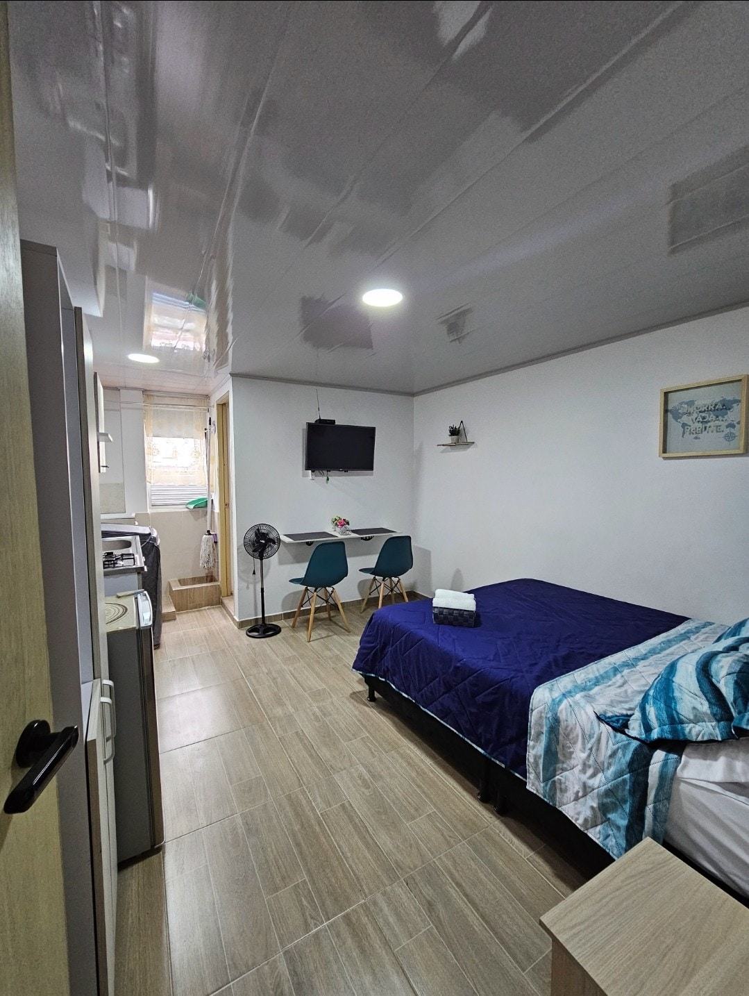 A well-lit room features a spacious bed covered with a blue blanket, a small dining table with two chairs, and a wall-mounted television. A fan is positioned nearby. Natural light enters through a window, and a kitchenette is visible in the background.