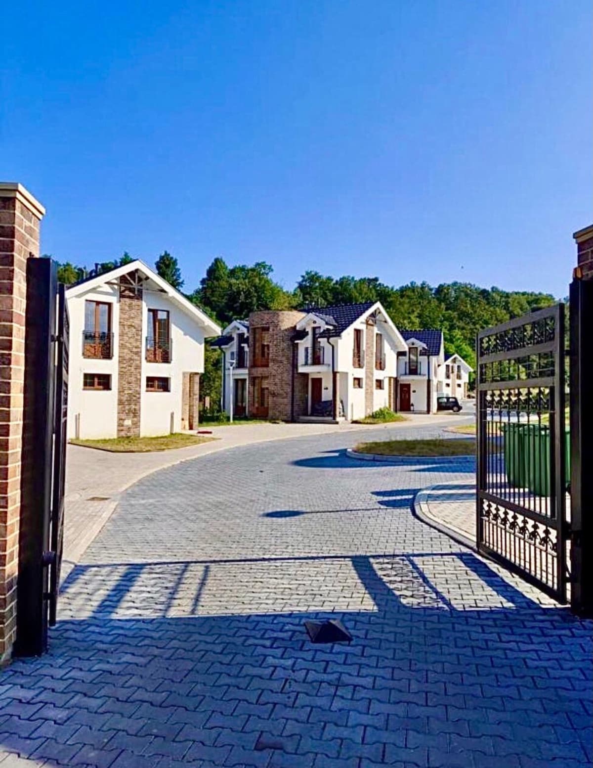 The entrance to the villa complex is framed by decorative gates, revealing a paved pathway that leads to several modern villas. Lush greenery is visible in the background, and clear blue skies enhance the inviting appearance of the neighborhood.