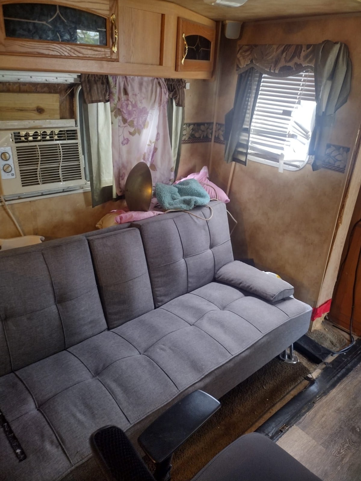 A gray fabric sofa bed is positioned by a window adorned with light curtains. A small pillow rests on the sofa, while a pink blanket and towel are casually draped nearby. An air conditioning unit is mounted on the wall above the seating area.