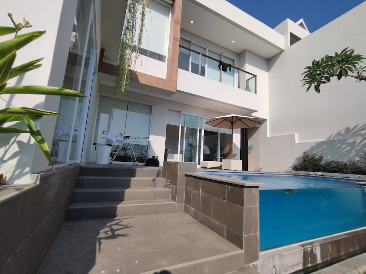 A modern exterior features a sleek design with large windows allowing natural light. Steps lead down to a pool area, where an umbrella provides shade. A small seating area is visible near the pool's edge, surrounded by greenery.