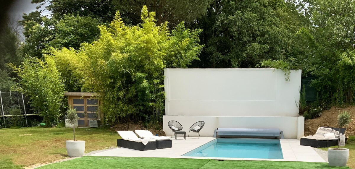 The outdoor area features a heated pool surrounded by a tiled deck. Two lounge beds are positioned for relaxation, and green landscaping adds a natural touch. A trampoline can be seen in the background, enhancing the recreational space.