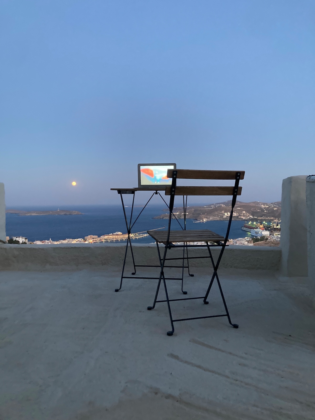 A small outdoor terrace features a sleek black table and a folding chair, overlooking a serene sea view. The table holds a laptop, with the backdrop of the city and the moon casting a soft glow across the horizon.