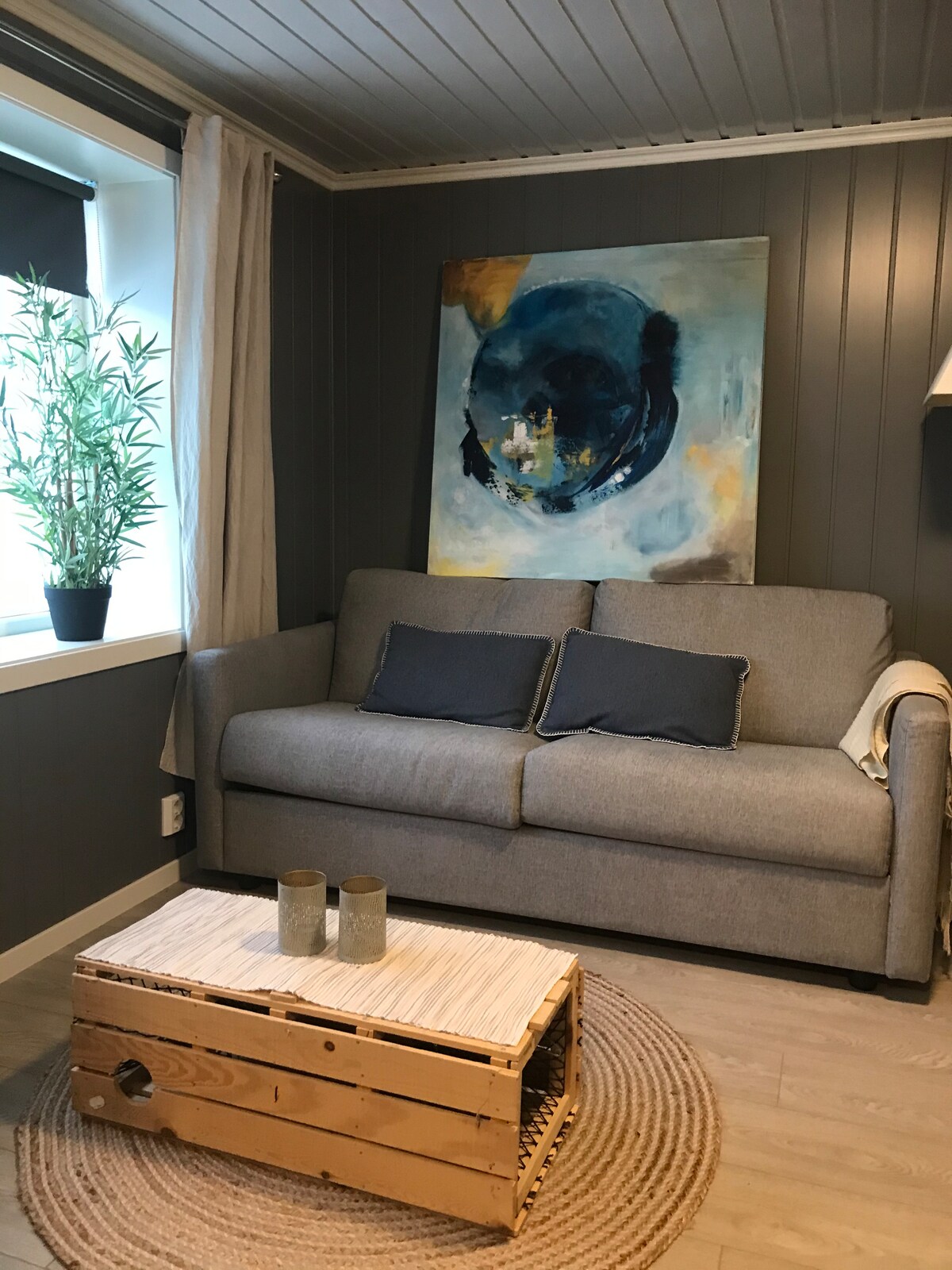 A cozy seating area features a grey sofa adorned with two decorative pillows. A colorful abstract painting resides on the dark wall above the sofa. A small wooden coffee table sits atop a circular woven rug, accompanied by two decorative candles. Natural light enters through a nearby window.