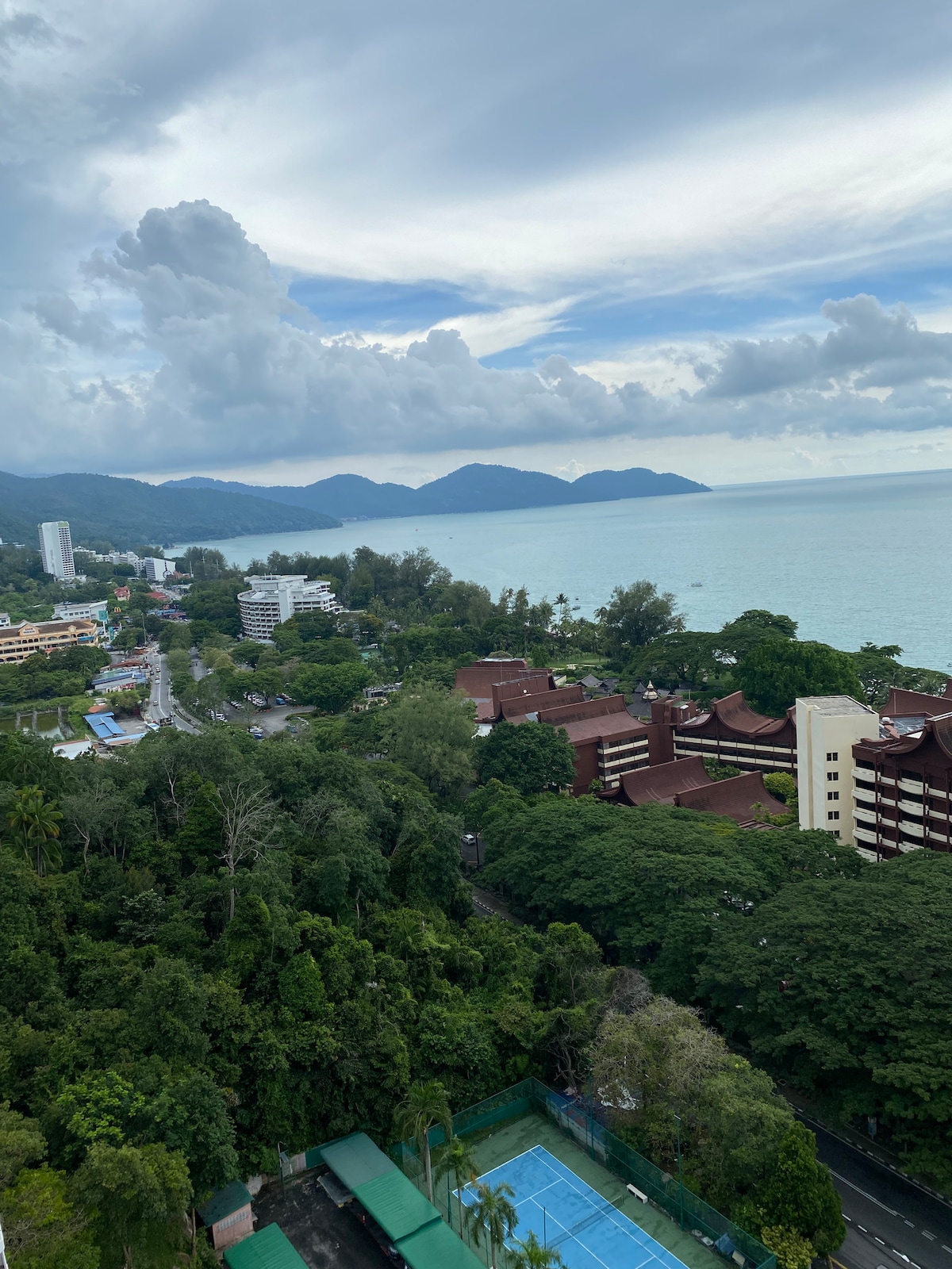 A panoramic view showcases the coastal landscape from an elevated position. Verdant trees frame the foreground, while the turquoise sea extends towards distant mountains under a partly cloudy sky. In the mid-ground, several residential buildings blend harmoniously with the lush surroundings.