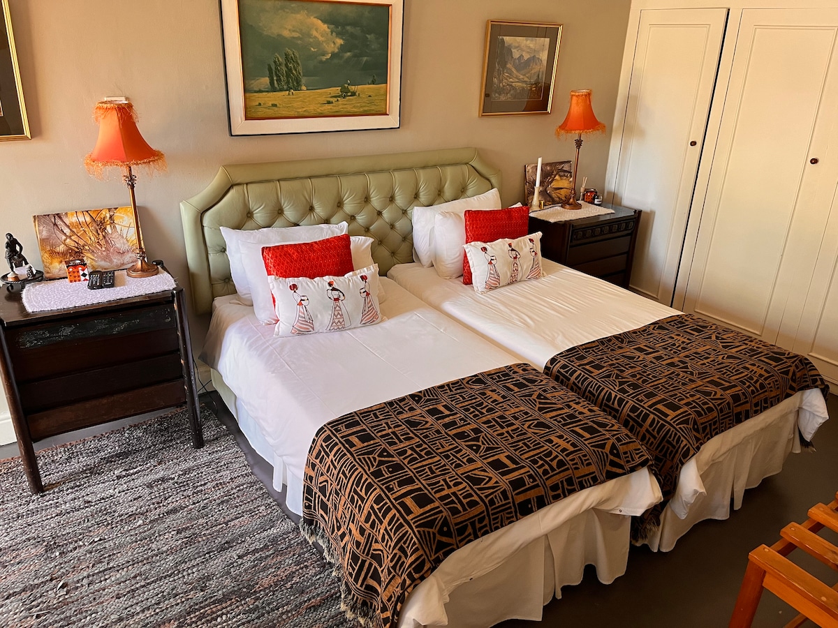 The room features two spacious beds, each adorned with decorative pillows and warm blankets, flanked by bedside tables with lamps. Artwork and scenic imagery decorate the walls, complemented by a wardrobe and a textured rug underfoot.