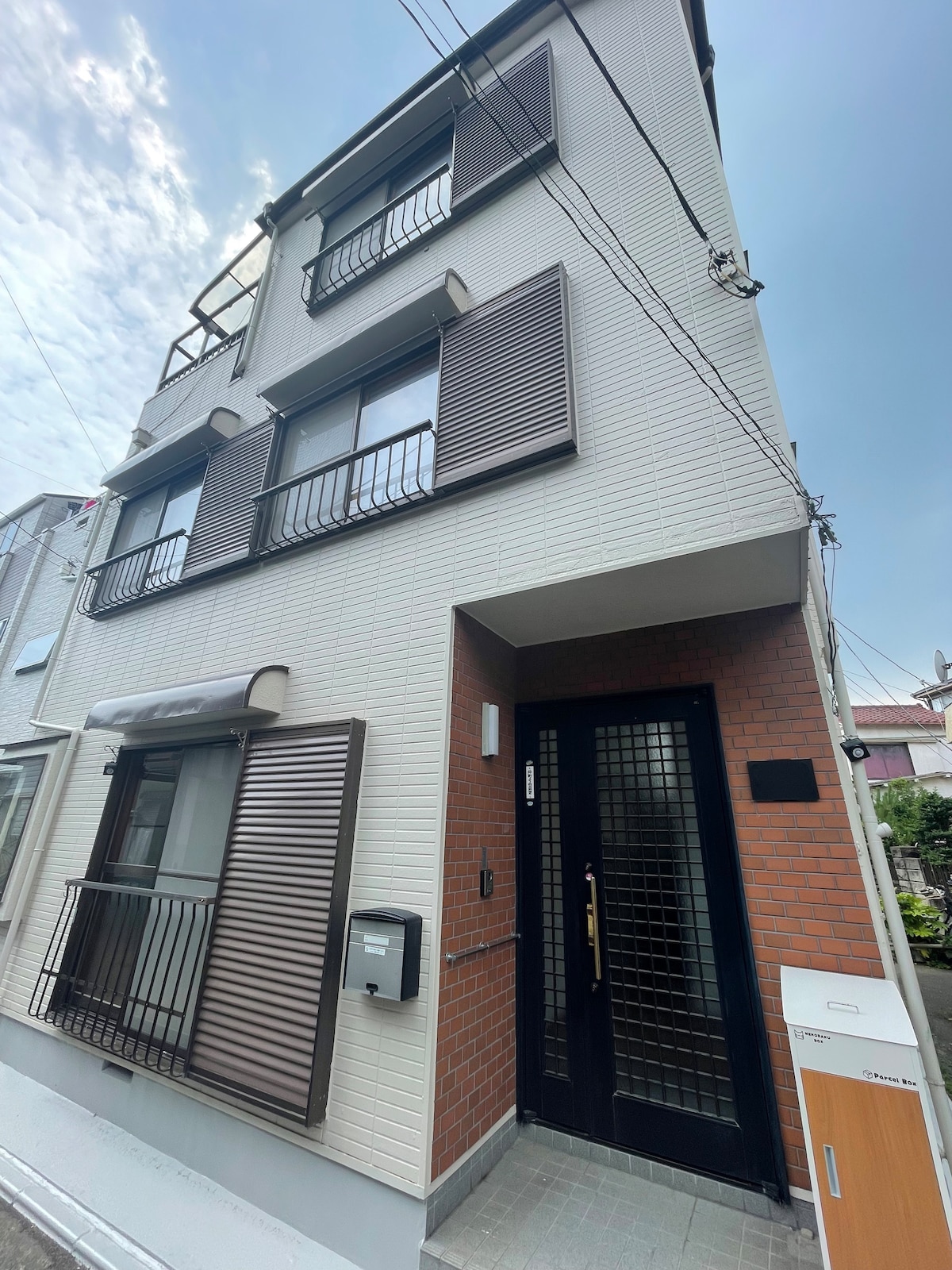 The exterior of the house features a modern design with a blend of light-colored brick and cream walls. The front entrance is marked by a black door with a grid pattern, accompanied by horizontal slatted shutters on the upper windows. A small mailbox is mounted beside the entrance.