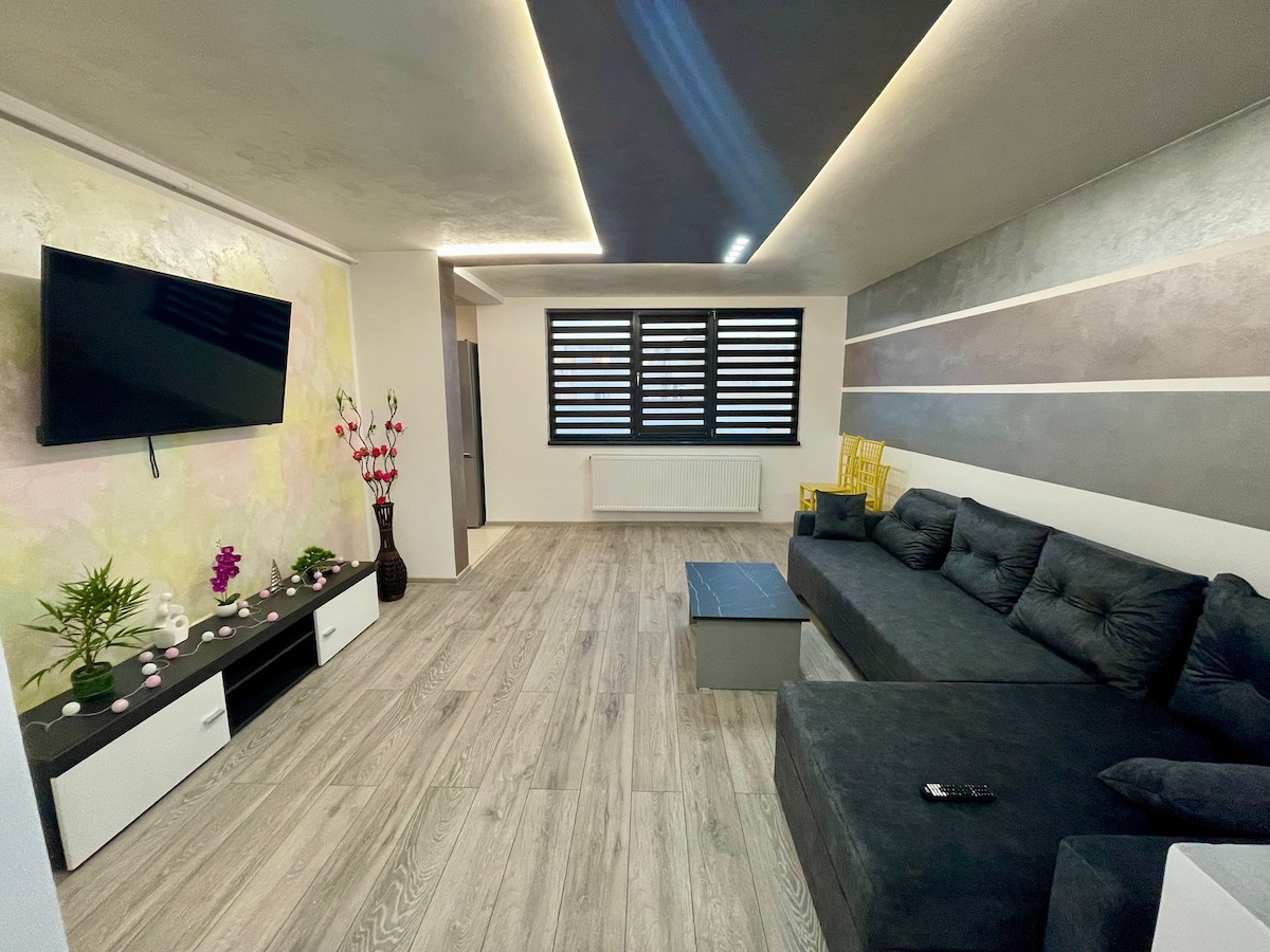 A modern living area features a spacious dark gray sectional sofa and a low coffee table. A wall-mounted television is visible, along with decorative plants and flowers. Horizontal stripes in neutral tones decorate the walls, complemented by shuttered windows that allow natural light to enter.