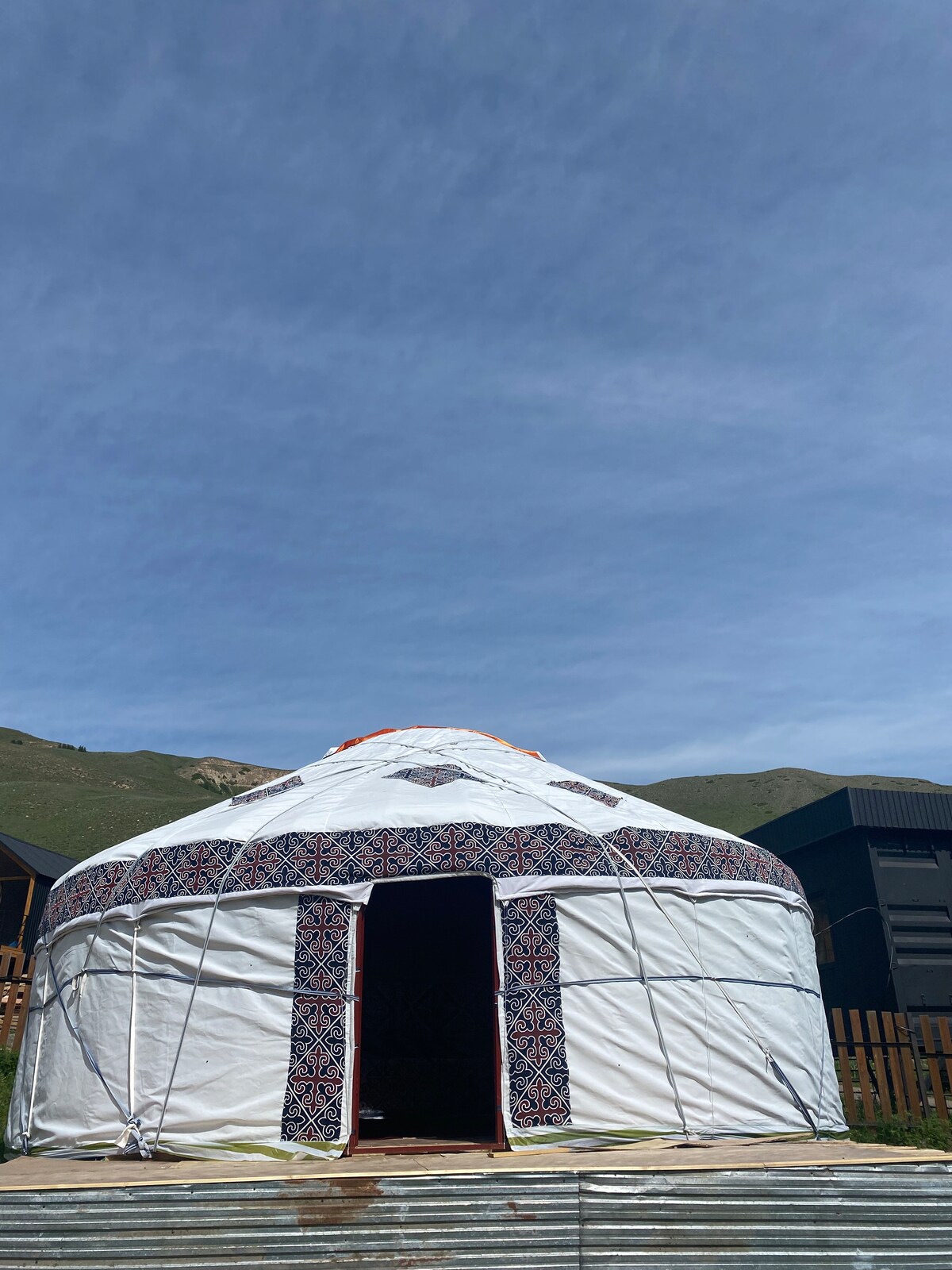 A traditional yurt is depicted with a white exterior and decorative patterns around the entrance. The surrounding landscape features rolling hills under a clear blue sky, adding to the tranquil setting. The structure stands on a wooden platform, inviting guests to explore.
